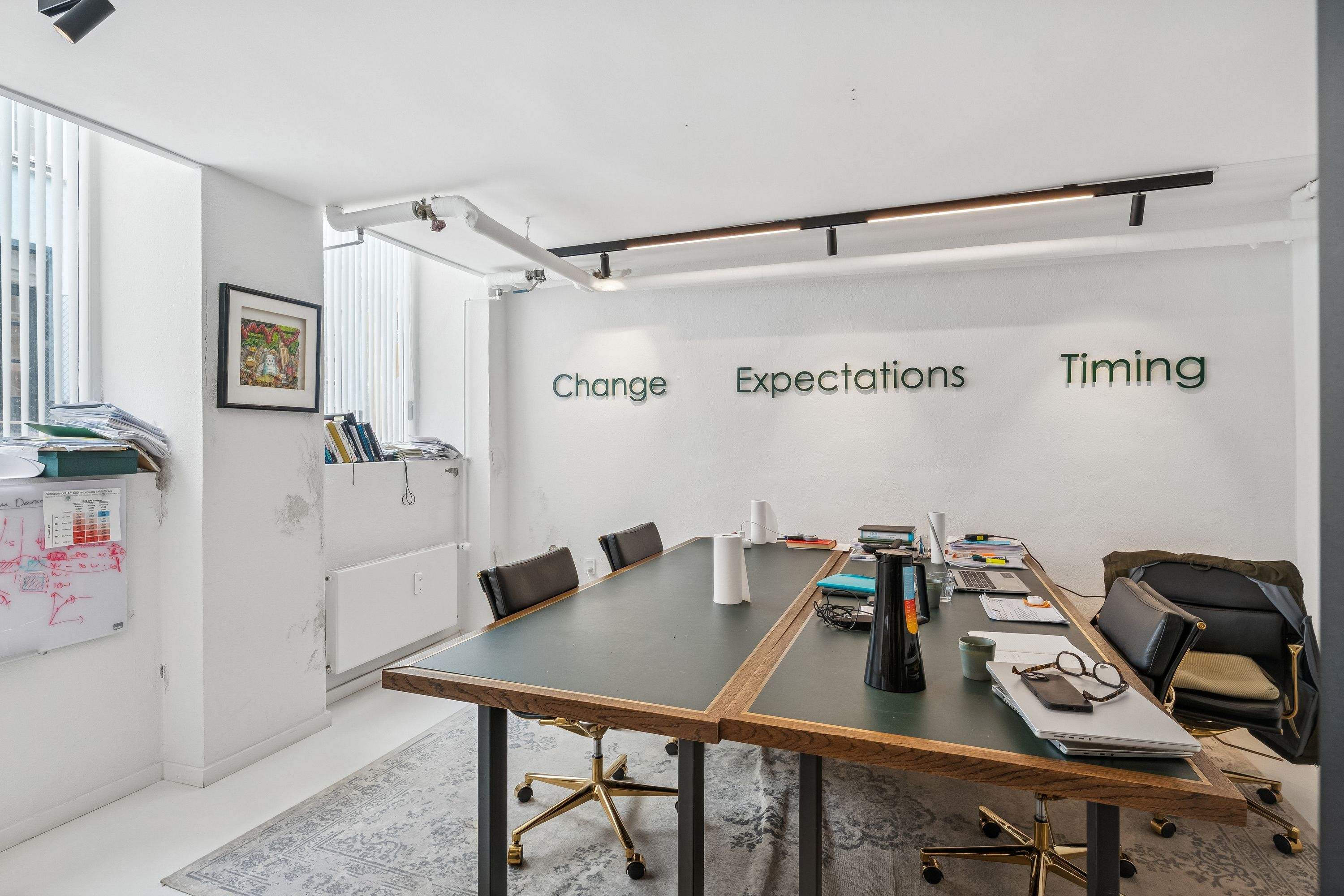 Kontor med bord og stole, skrivebord, bøger og møbler, gulvtæppe, hvide vægge, møbler med hjul, billede på væggen og indretning med tekster