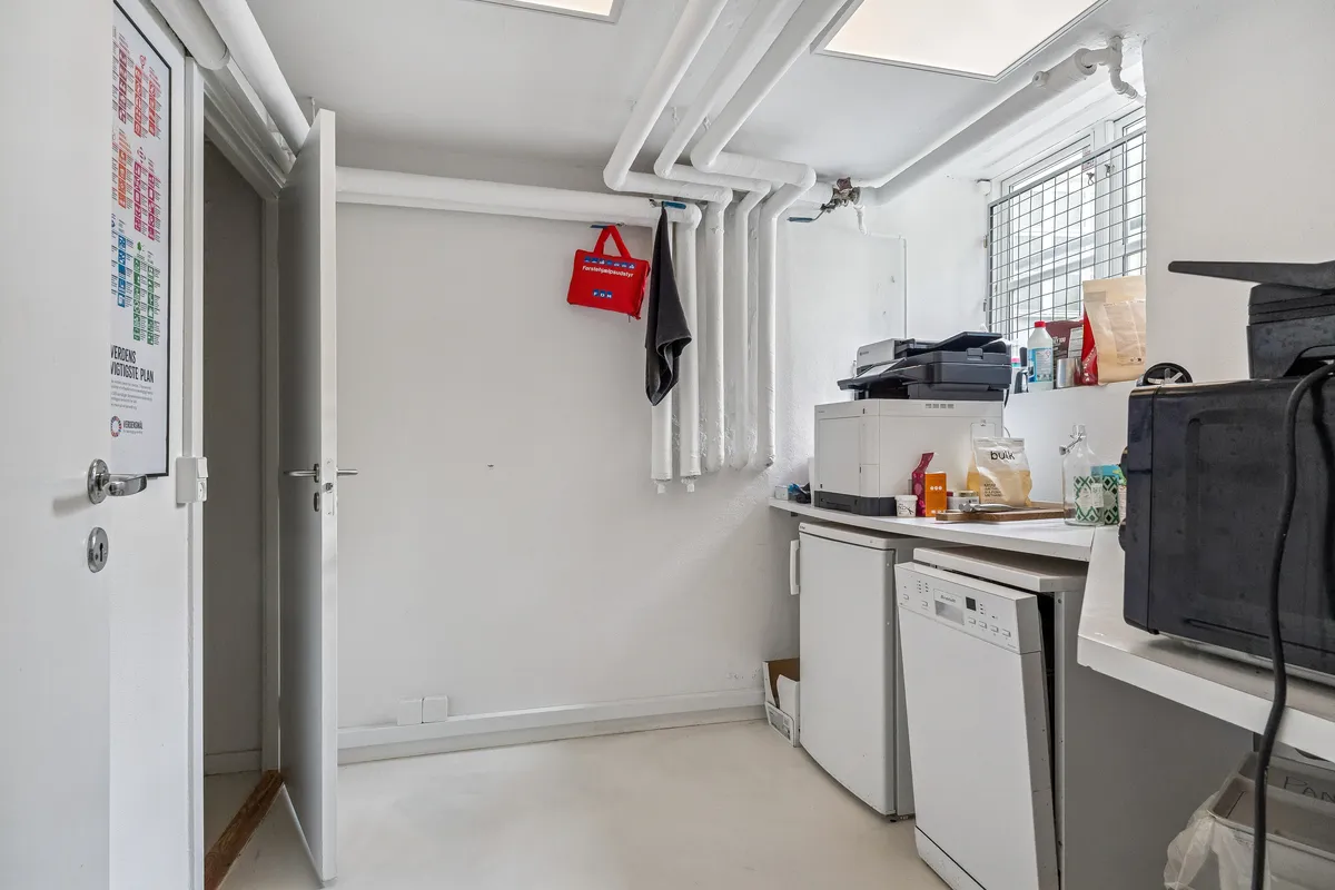 Rummelig kælderlejlighed med lyse lofter, møbleret køkken og moderne apparater samt en printer og flere ting på hylden.