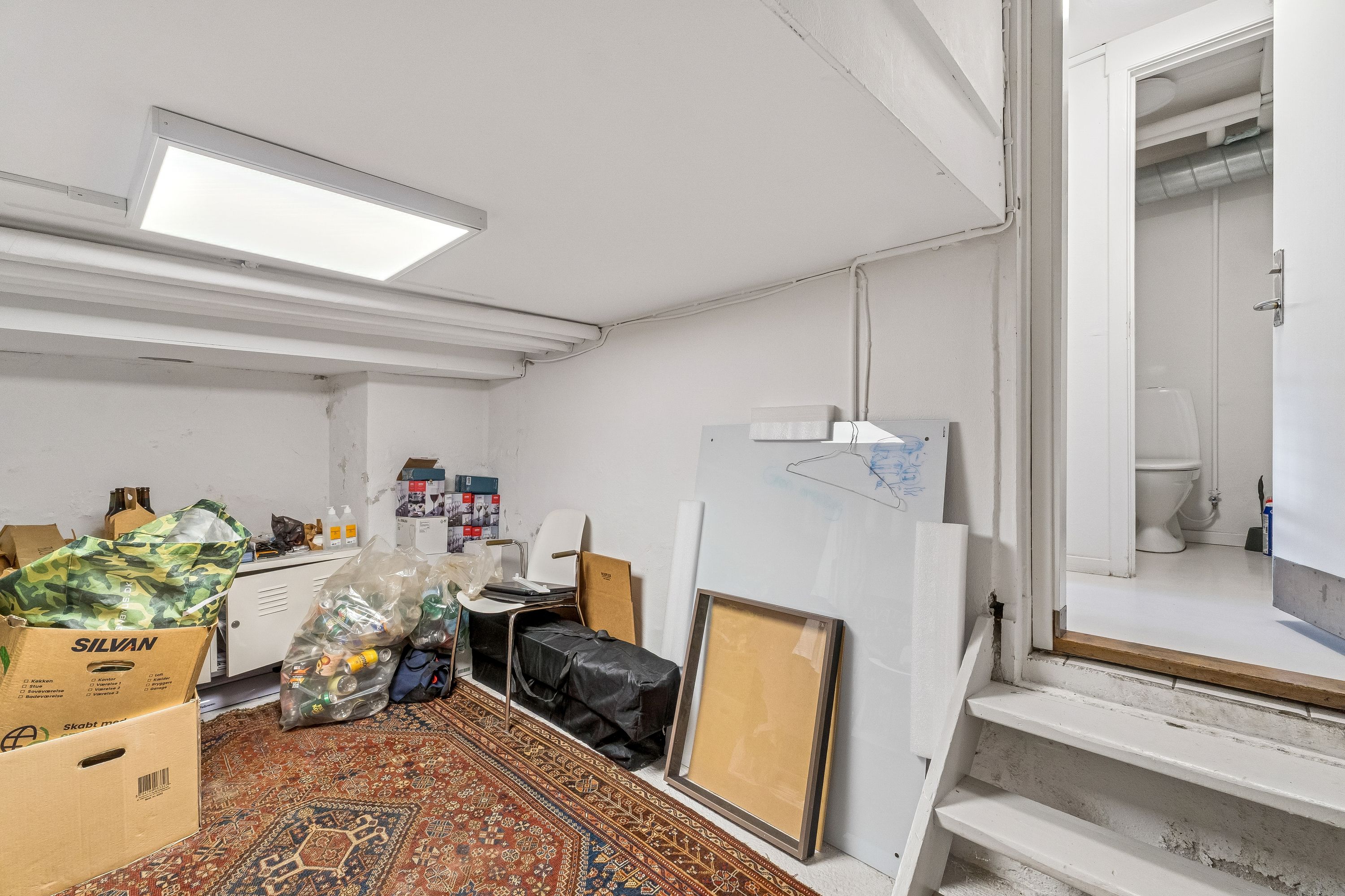 Erhvervslejemål på 72 m² med lyse lofter og et luksuriøst gulv. Rummet med lagerkasser, en hvid toilet og et stort hængemaleri.