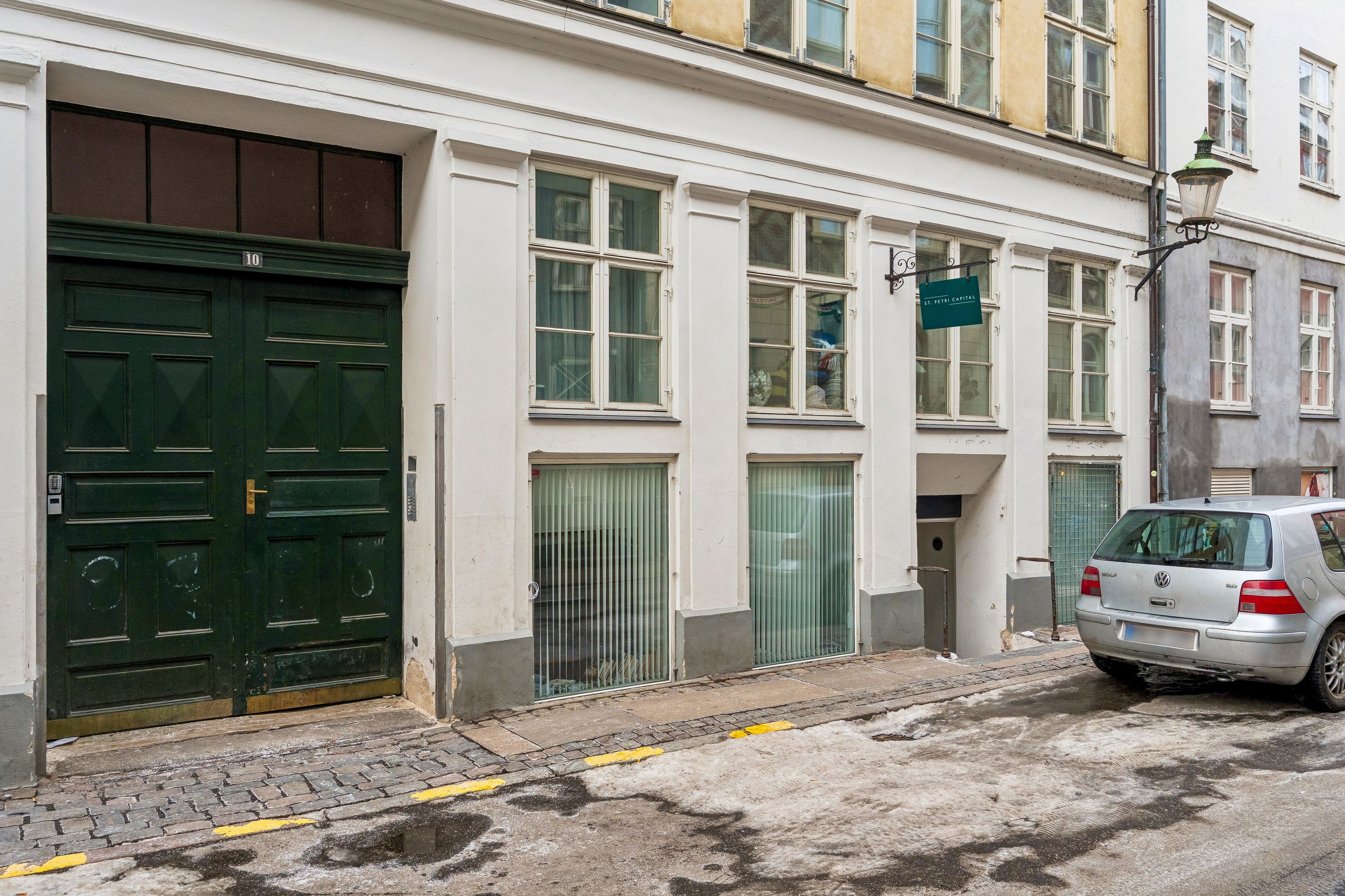 En gammel, hvid bygning med glasruder og en grøn dør i København. St. Petri Capital-skilt hænger på bygningen. En gul bil er parkeret foran bygningen.