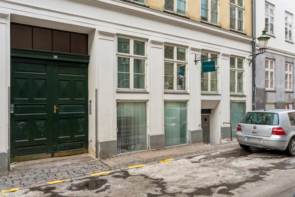 En gammel, hvid bygning med glasruder og en grøn dør i København. St. Petri Capital-skilt hænger på bygningen. En gul bil er parkeret foran bygningen.
