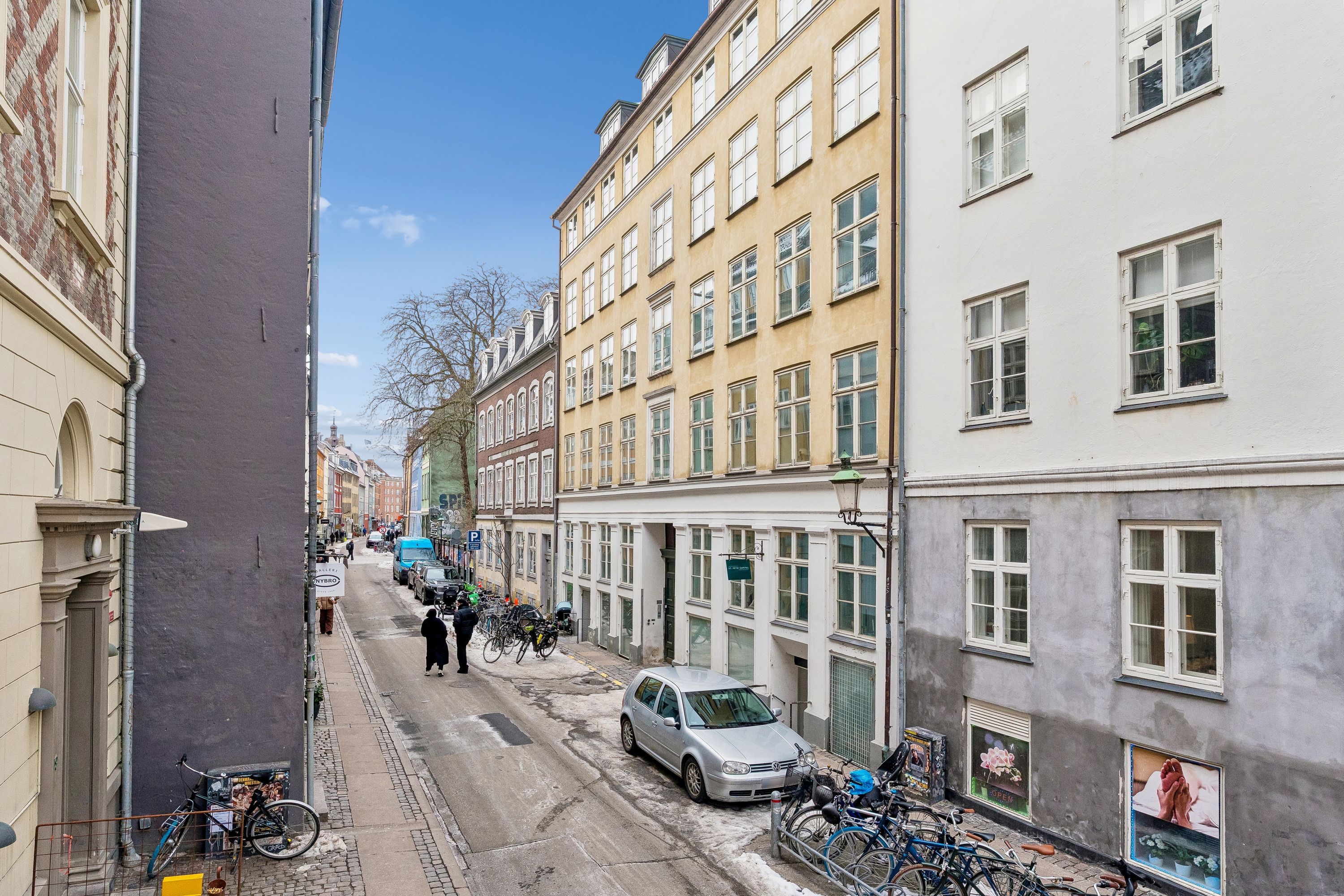 En smal gade med parkerede biler og cykler, hvor folk går langs siderne. Bygninger med mange vinduer og skrånende gavle. En parkeringsplads med flere parkerede biler og cykler.