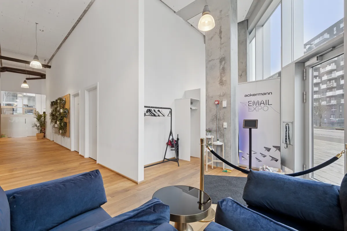 Moderne og rummeligt kontorlejemål med højt til loftet, lyse vinduer og naturligt lys. Pågulv er der trægulv og blå sofaer. Der er et skrivebord med papirfly og en banner med teksten "ackermann EMAIL EXPO".