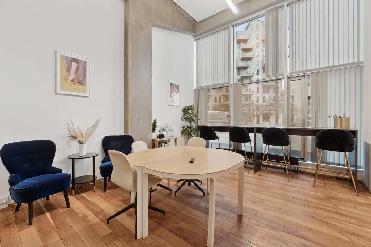 Kontorlejemål med moderne interiør, trægulv, ovalt bord og høje, komfortable stole. Vægdekorationer med lyse og moderne farver. Store vinduer med udsigt til bygninger udenfor.