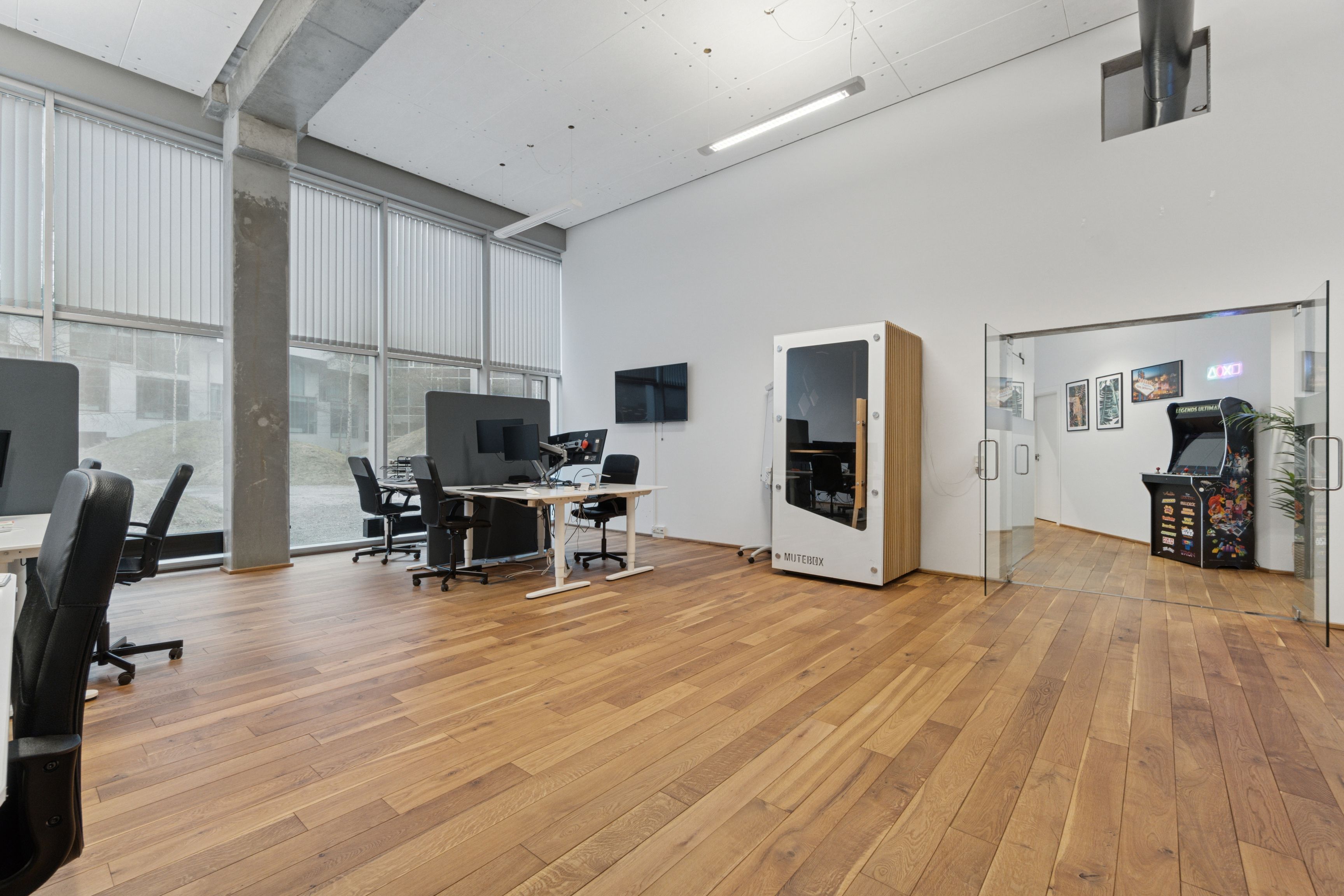 Kontorrum med lyse gulve, høje ruder, skriveborde, stole, monitorer, arkademaskine og lysekrone. Moderne og rummeligt lejemål i Ørestad med en åben glasdør og store vinduer