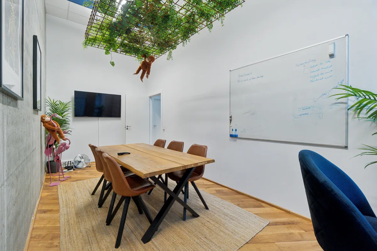 Kontorrum med mødebord, to flamingoer og en skabning, skrivebord, tv, og hængende grønne planter i loftet