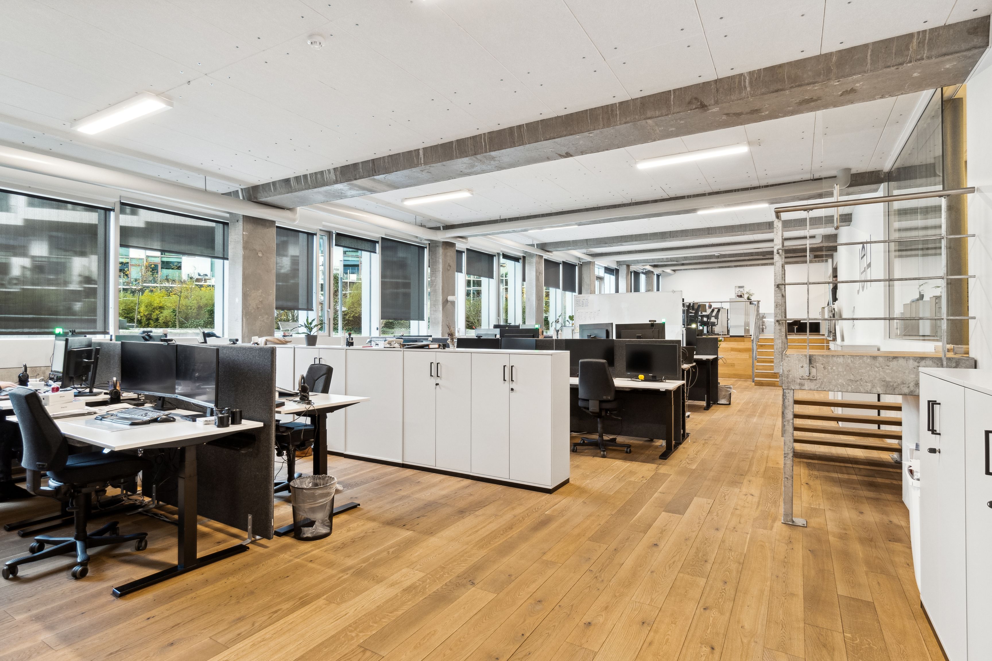 Kontorlejemål med lyse gulve, lysevinduer og moderne kontormøbler med computere og skriveborde. Forskud på lys og indbydende arbejdsmiljø med højt til loftet og metaltrapper.