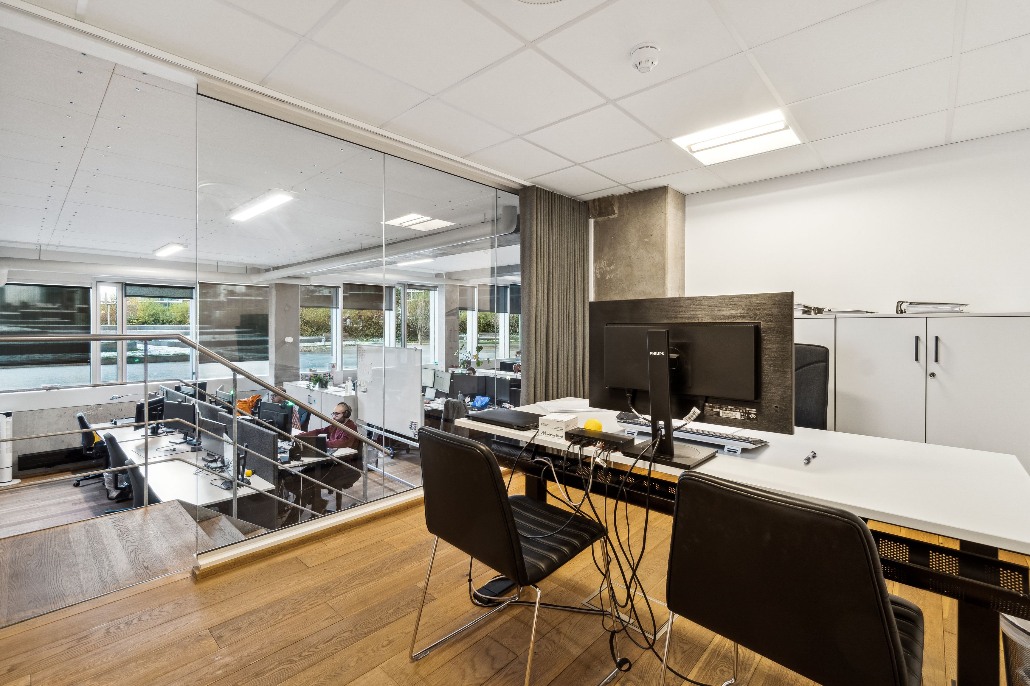 Lyst og moderne kontor med glasvægge, højloft, hårdt trægulv, skrivebord, computer, stol, skriftbord, sofa og mange vinduer med udsigt til grønne træer