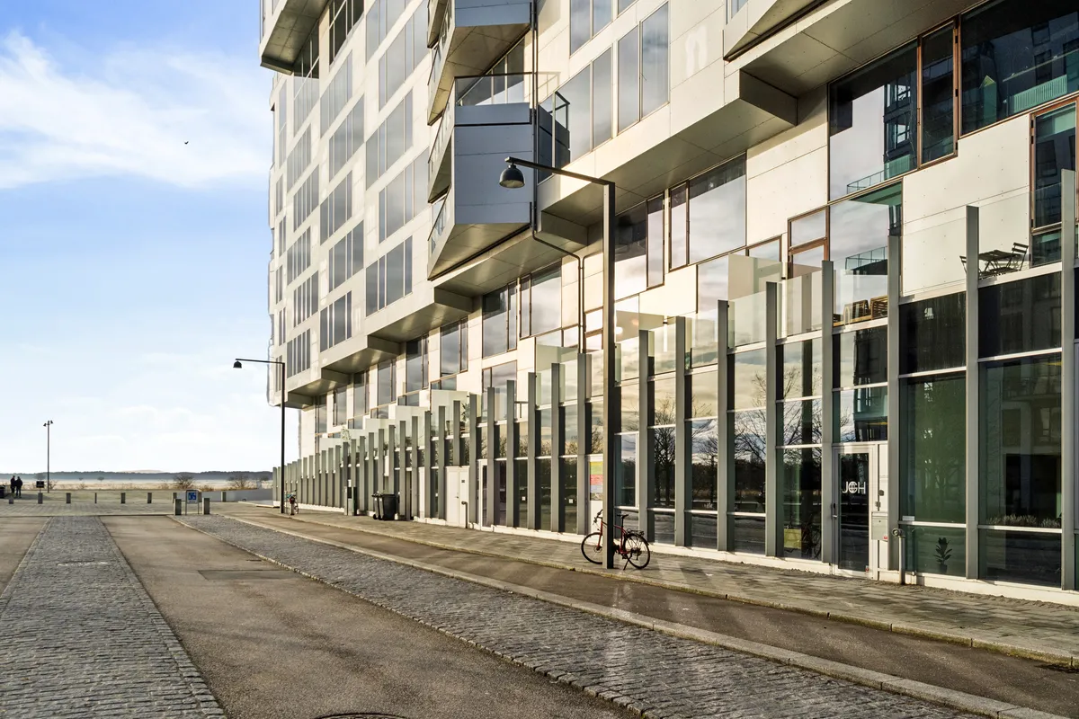Moderne og lyst kontorlejemål med glasvinduer og balkoner langs Ørestad. En cykel står foran bygningen, og der er en søjle med et lys på gaden. Baggrund med bjerge og himmel med skyer.