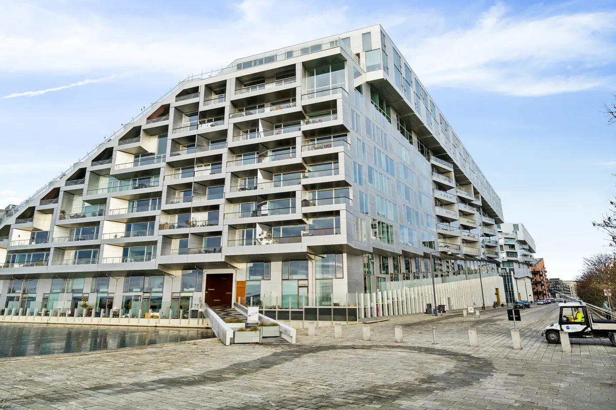 Moderne højhus med glasvinduer og balkoner ligger ved vandet. Højhus med hjørneplacering og terrasse på toppen. Moderne kontorlejemål med mange parkeringspladser og biler parkeret.