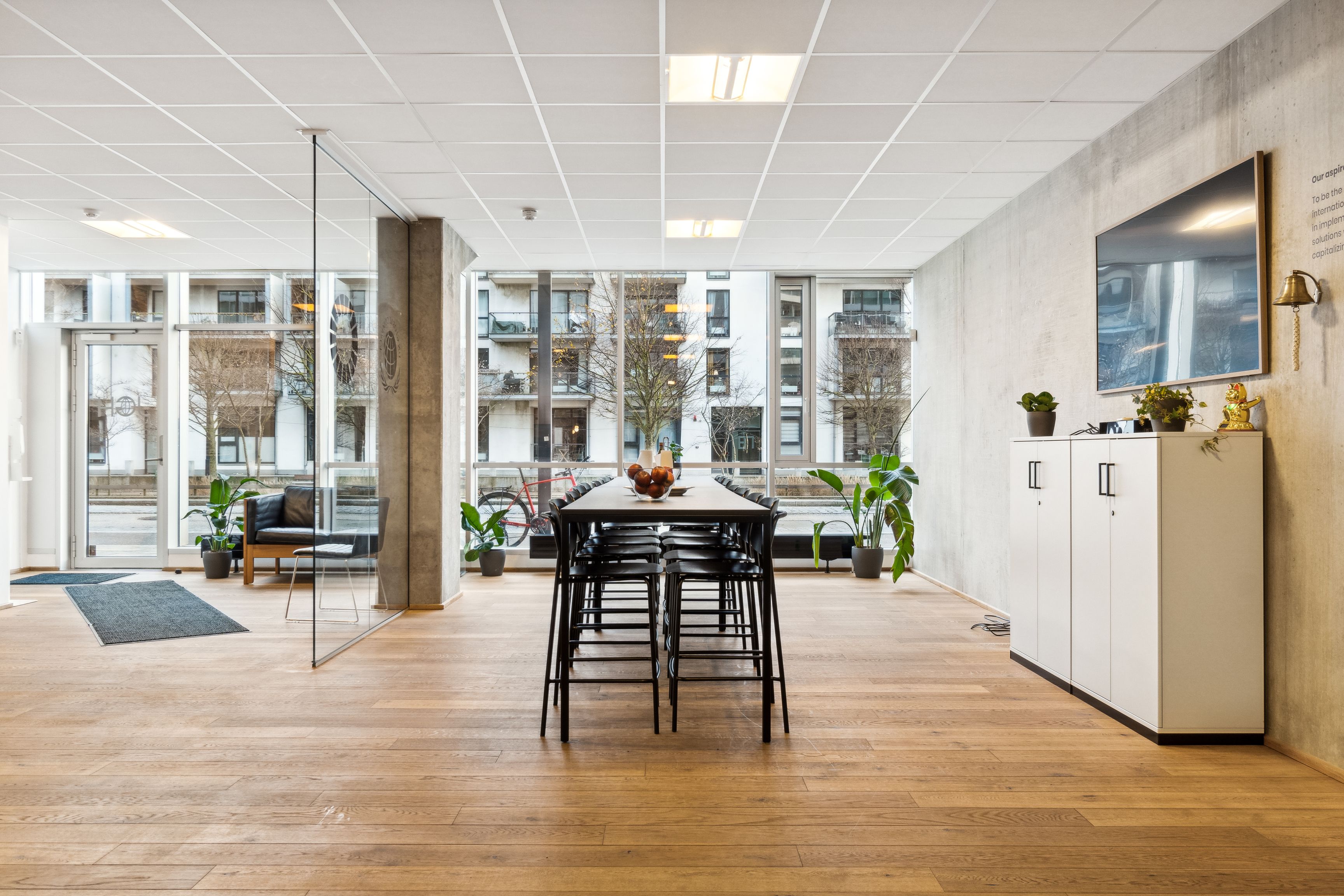 Lyst kontorlokal i Ørestad med lyse vinduer og trægulv, moderne møbler, plantepotter og en stor tv-skærm på væggen