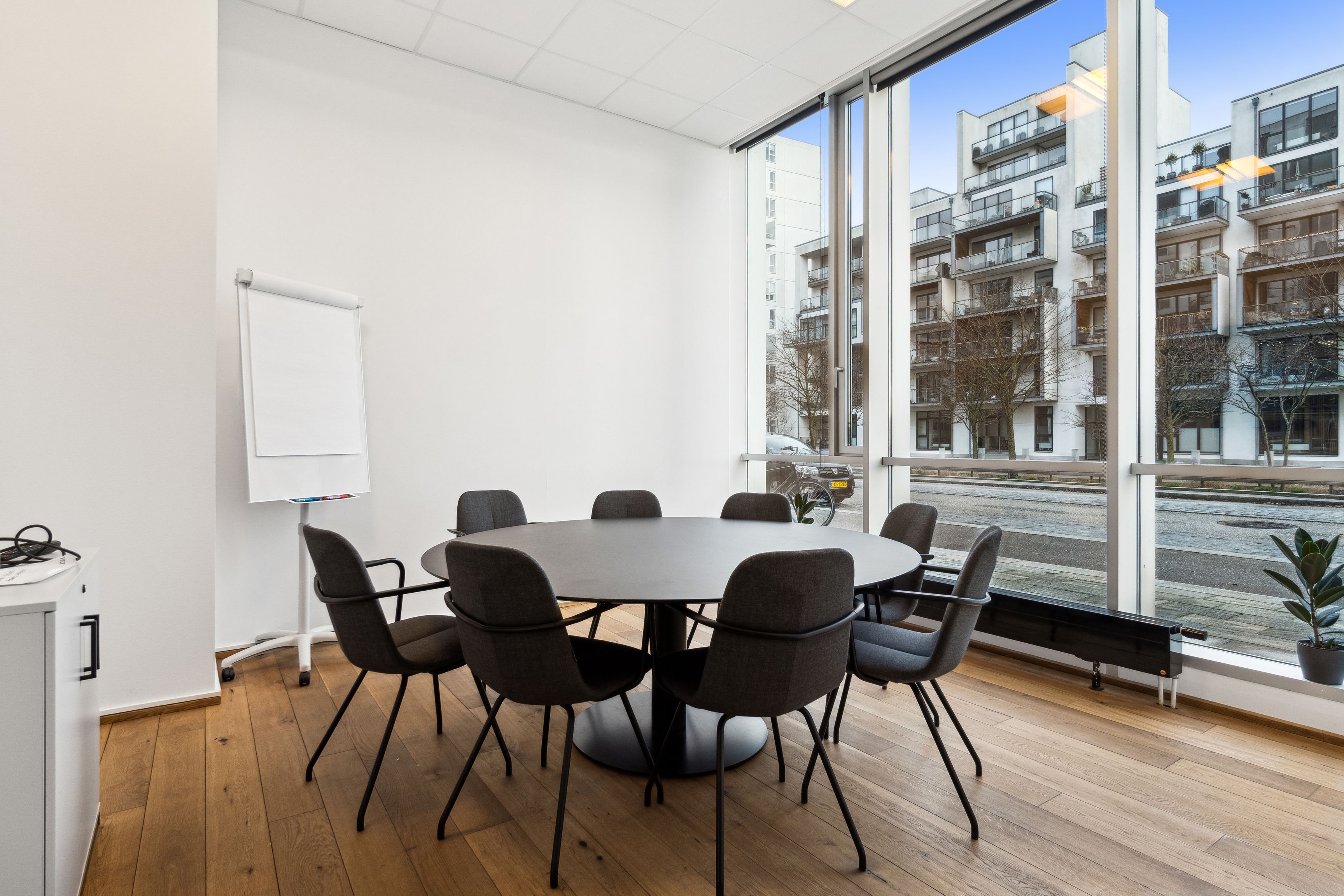 Kontorlokaler med store ruder og runde bord, højloftet, lyse og moderne møbler, hvid væg og gulv, hvidt tavlebord og plante i vinduet