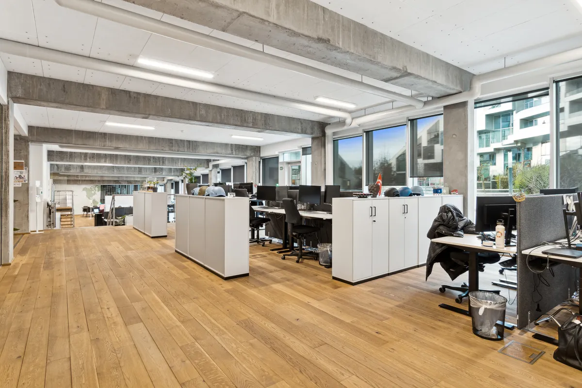 Kontor med lyse gulve, lyse lofter, store vinduer, højteknologiske skriveborde og møbler. I midten er der en række skriveborde og stole. I baggrunden er der en bygning med terrasser og en skov.