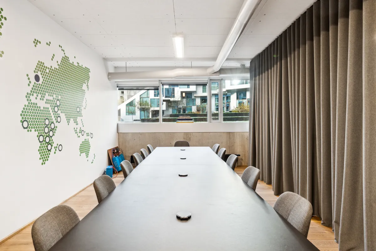 Lyst og indbydende kontor med lyse gulve og hvide vægge med grønt punktmønster. Langt borde med ståloverflade og læderstole. Solskin trænger ind gennem store vinduer med udsigt til bygninger.