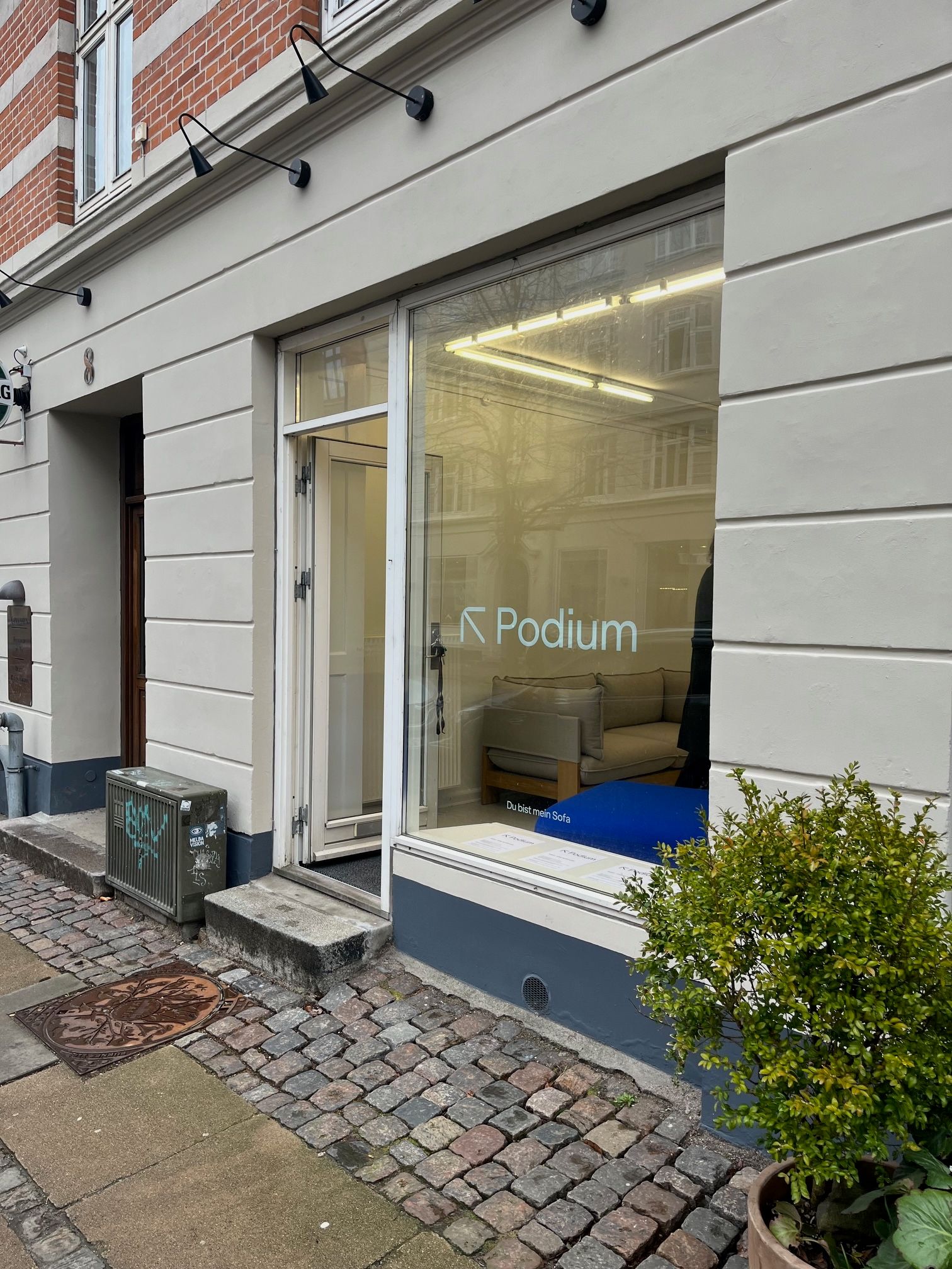 Butiksvinduet til leje på Århusgade 8 viser et sovesofa og udsigt til en skov. Der er plante i urtepotte og en åben dør.