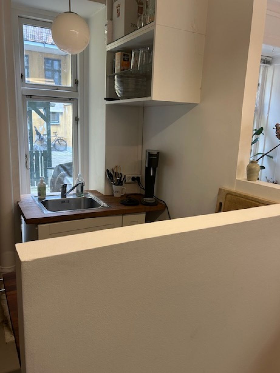 I en ren og moderne køkkenafdeling med en hvid væg og lyse loftslamper er der et træbord og et håndvask. Et køkkenapparat står på gulvet.