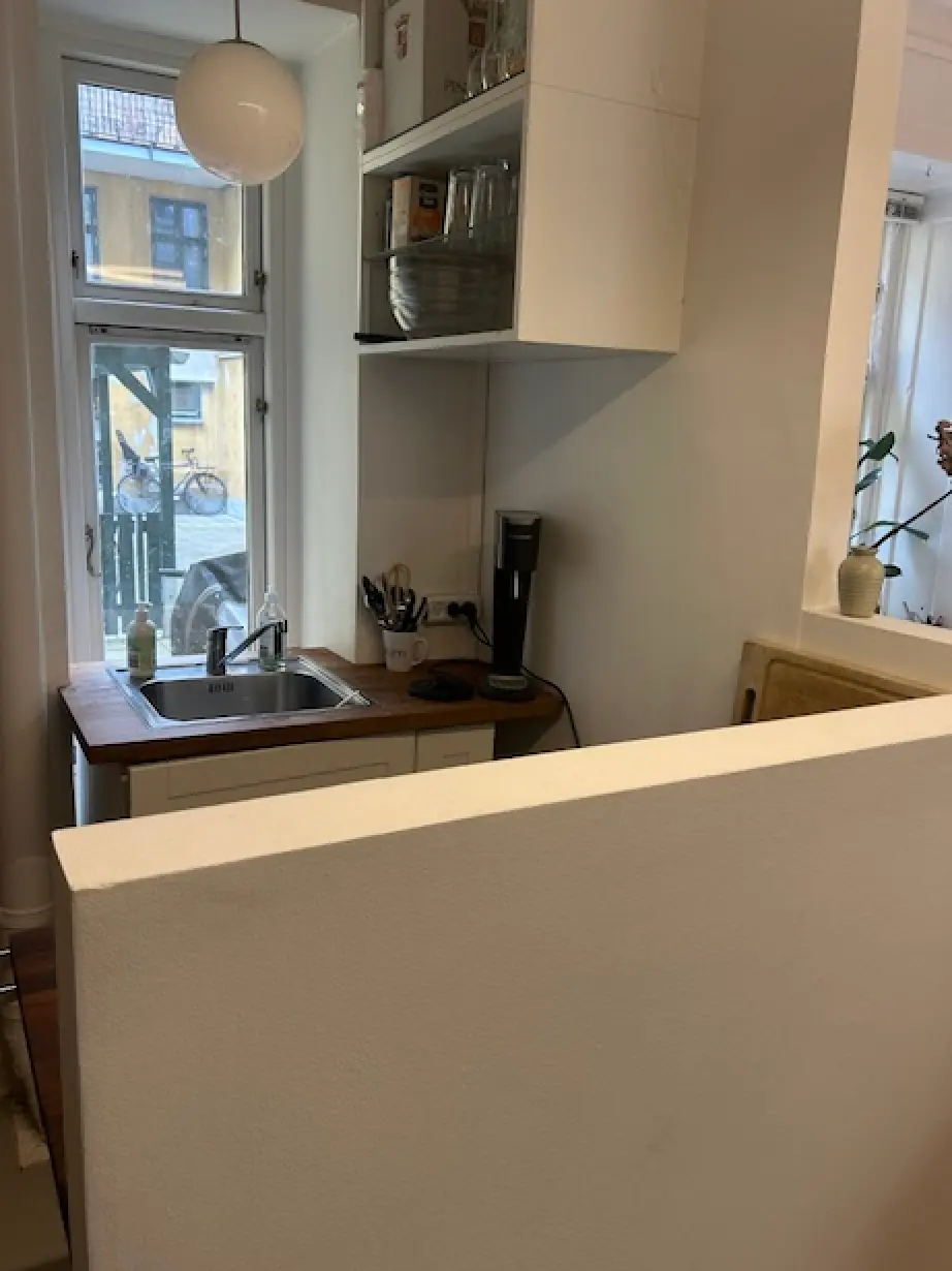 I en ren og moderne køkkenafdeling med en hvid væg og lyse loftslamper er der et træbord og et håndvask. Et køkkenapparat står på gulvet.