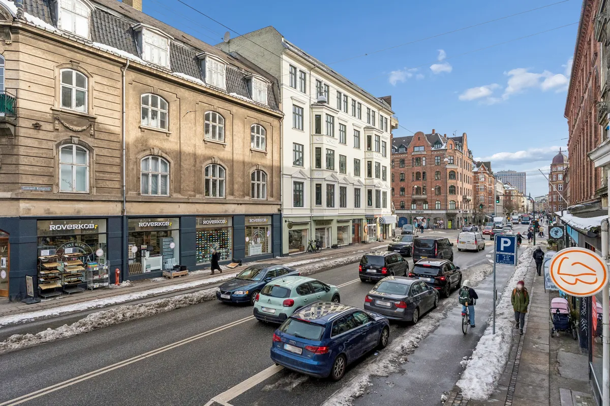 Personer går langs fortovskanterne og biler er parkeret langs vejen på en gade i Frederiksberg med butikker og bygninger langs vejen