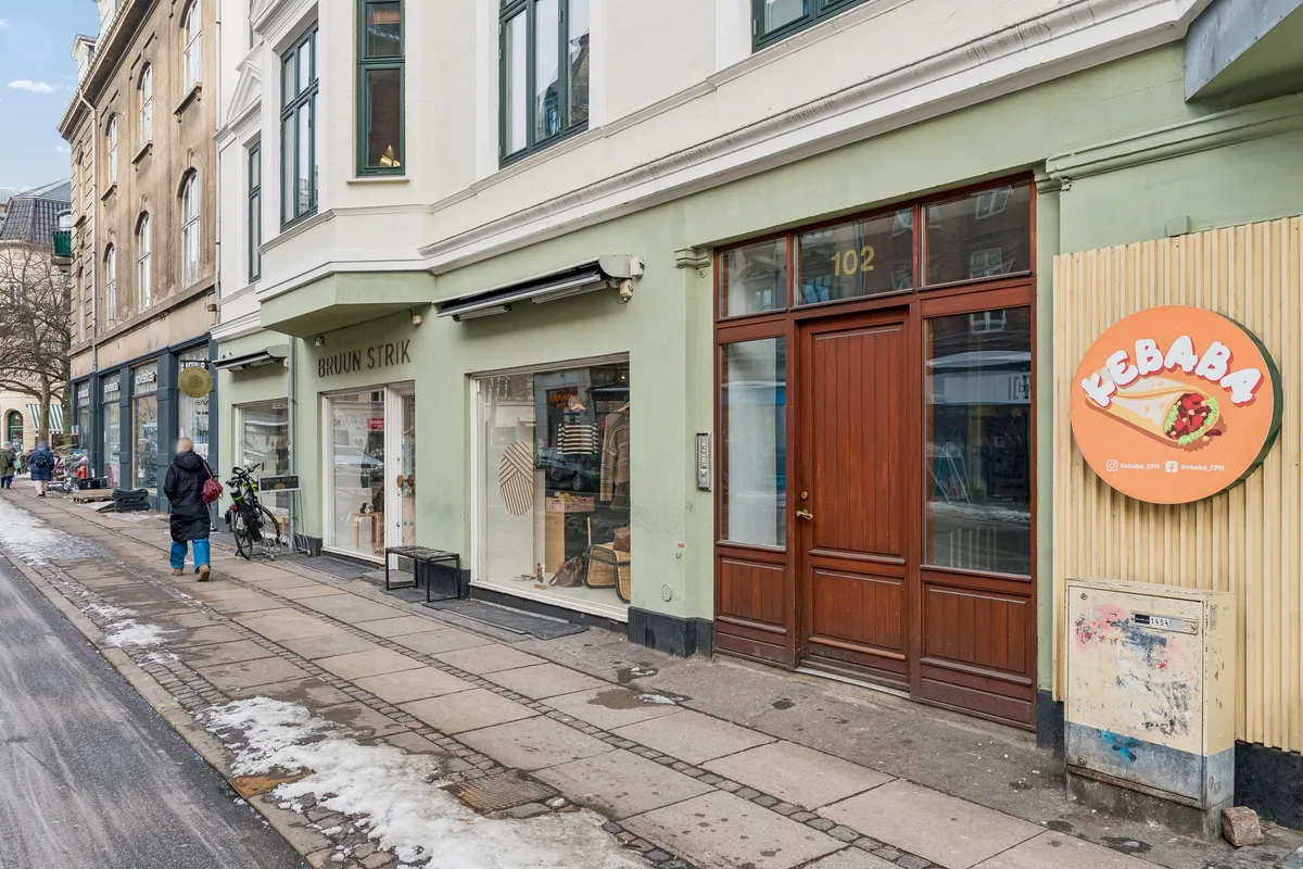 Kontor til leje på Gammel Kongevej 102, 1. tv, 1850 Frederiksberg C. Butikslokaler med glasvinduer og skilte på grønne facader. En person går forbi på fodgængerbroen med sne.