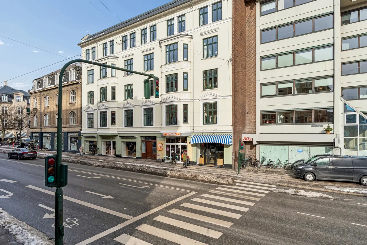 Kontorbyggeri og gågade med cykler og biler i forgrunden og en grøn trafiklygte i midten af gaden og en blå-hvid pavillon foran en af bygningerne