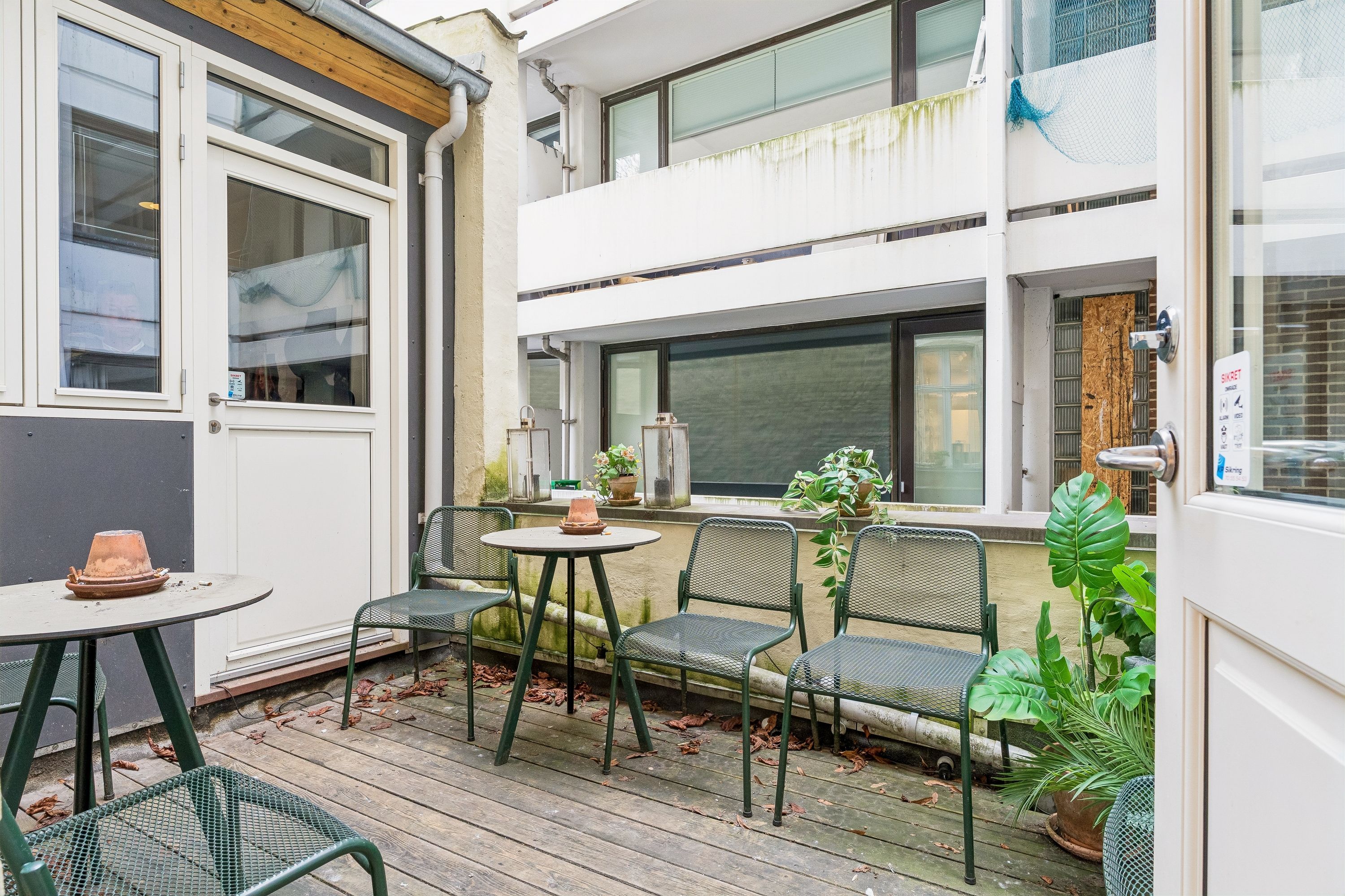 En have med bord, stole og planter. En åben dør med tekst på det. Et glas vindue med et plantepotte og en lampeskærm. Et lysthus med et loftsvindue og en skorsten.