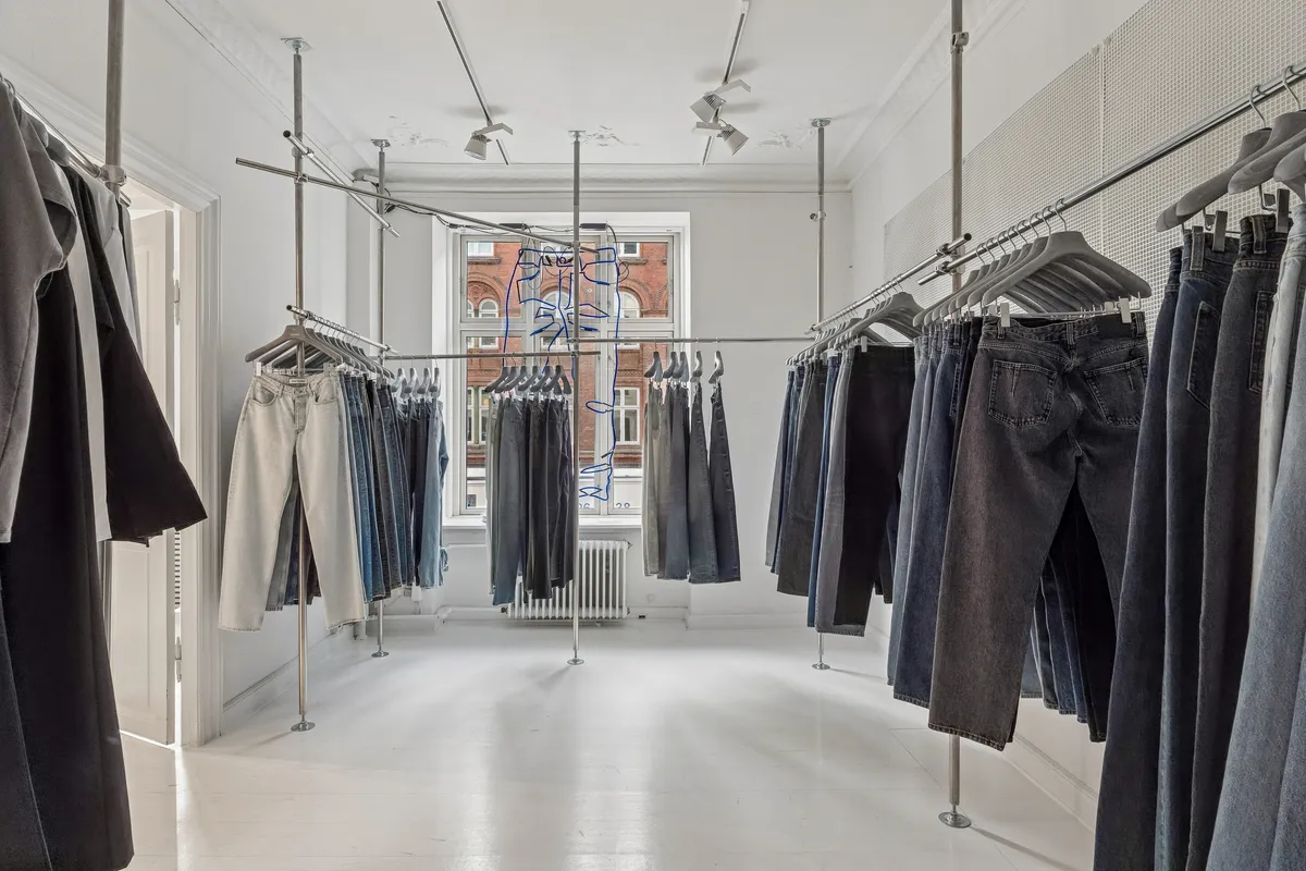 Sorte og hvide bukser, samt andre tøj hængende på klædhængere og metalstandere i en rummelig, hvid og minimalistisk kontorlokal.