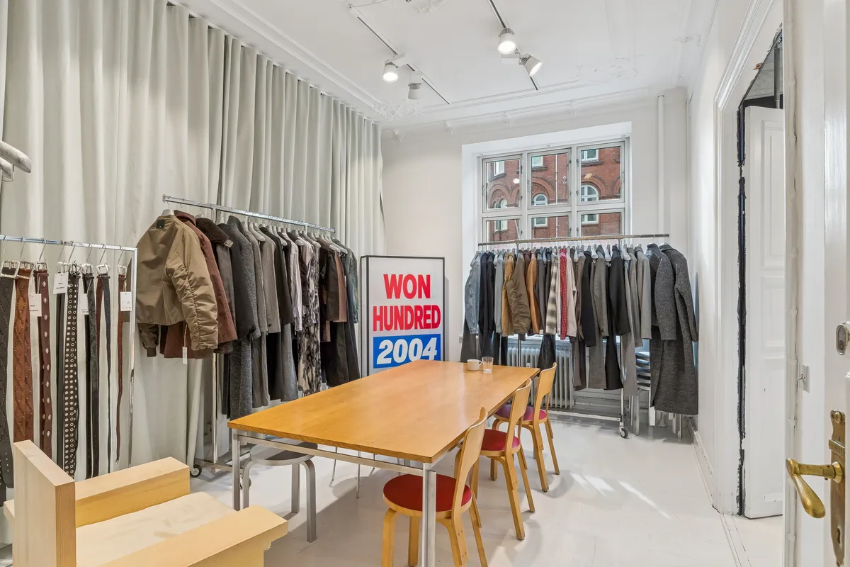 Kontor til leje med lyse hvide vægge, lysekroner og træbord med stole og træskabe med klæder i baggrunden.