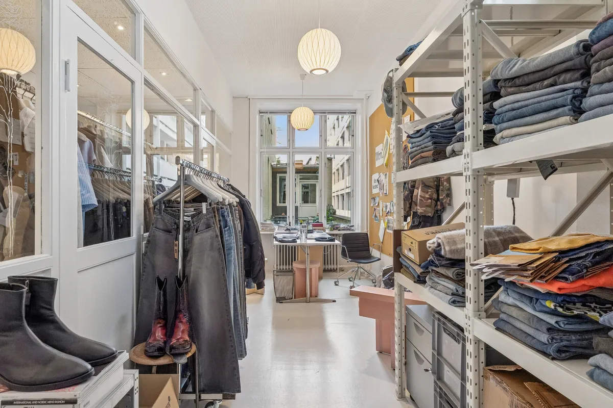 Kontor med lyse gulvvægge og moderne design. Sorte støvler og jeans på hjulstativ og høj hylde med foldede jeans og trøjer. Bord med papirkurv og skrivebord i baggrunden