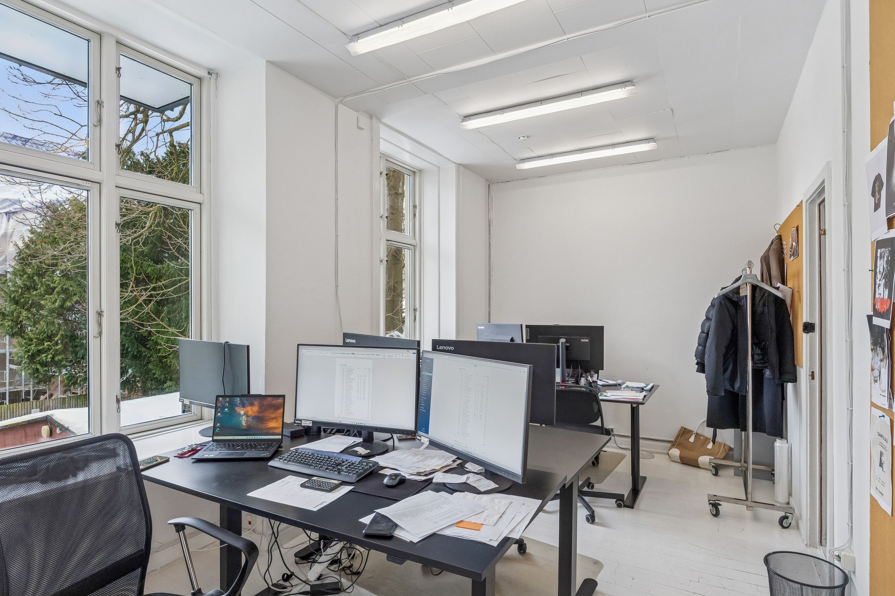 Kontor med lyse gulvvedligeholdelse og vinduer med udsigt til grønne træer og lyse himmel. Moderne møbler, computere og papir fordelt på et bord.