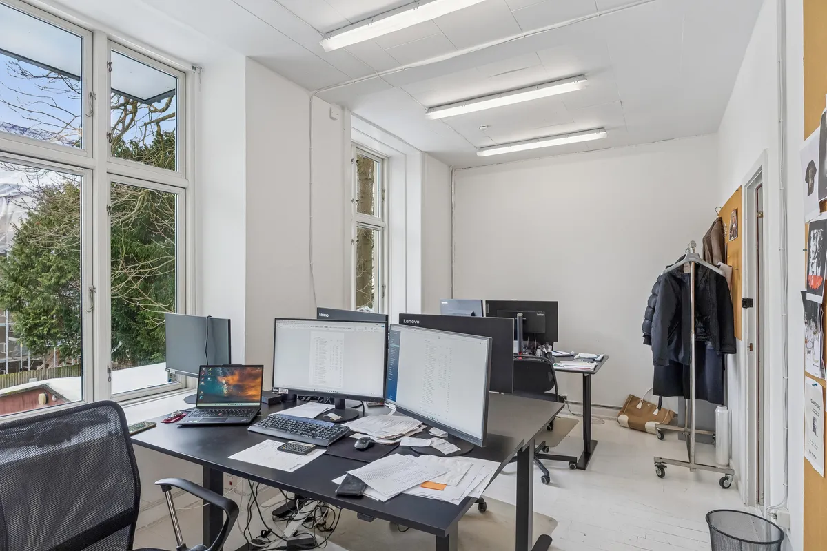 Kontor med lyse gulvvedligeholdelse og vinduer med udsigt til grønne træer og lyse himmel. Moderne møbler, computere og papir fordelt på et bord.