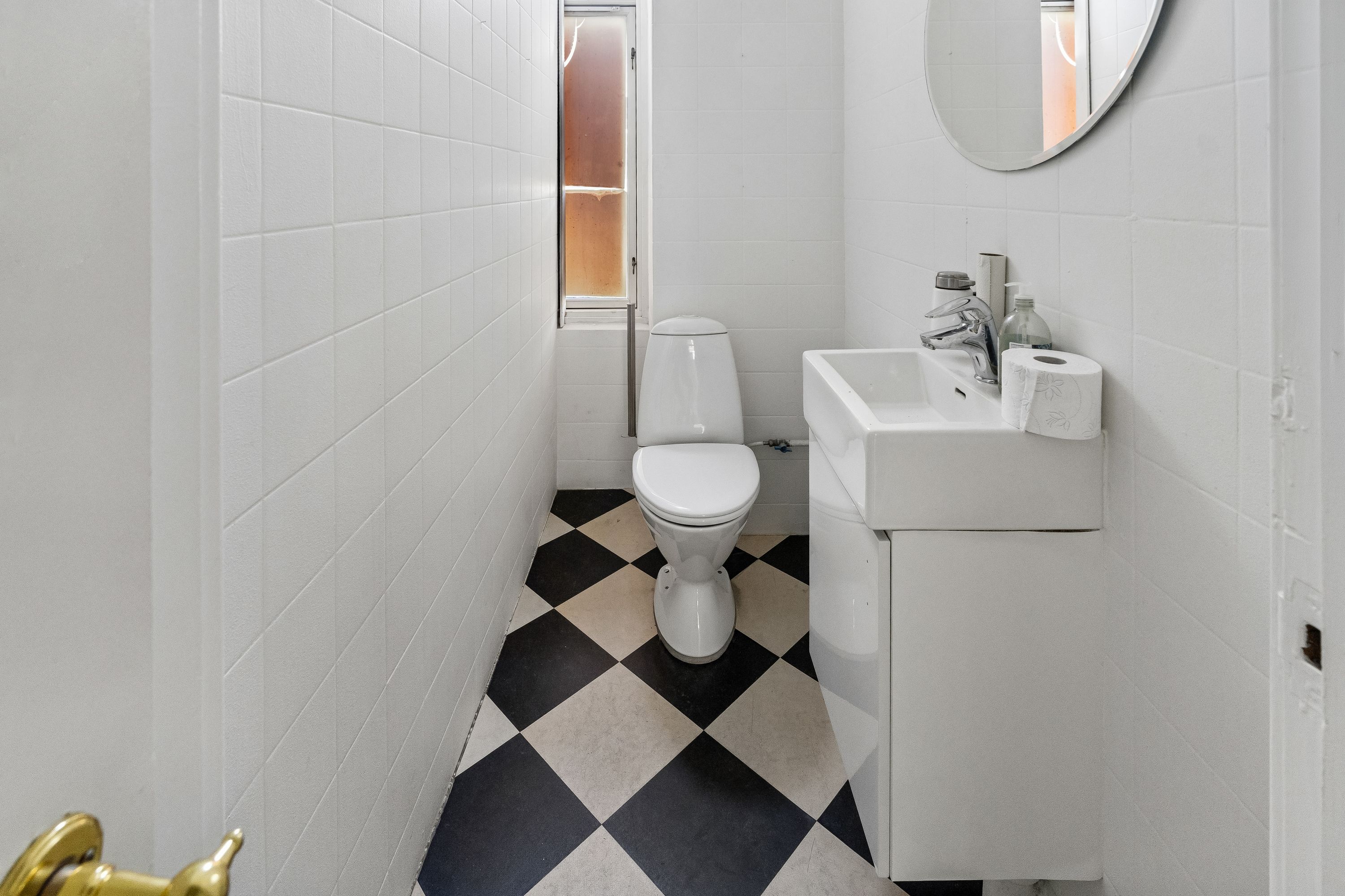 En lille badeværelse med hvidt flisede vægge og gulv med sort og hvidt tæppe og hvidt toilet og hvidt vask.