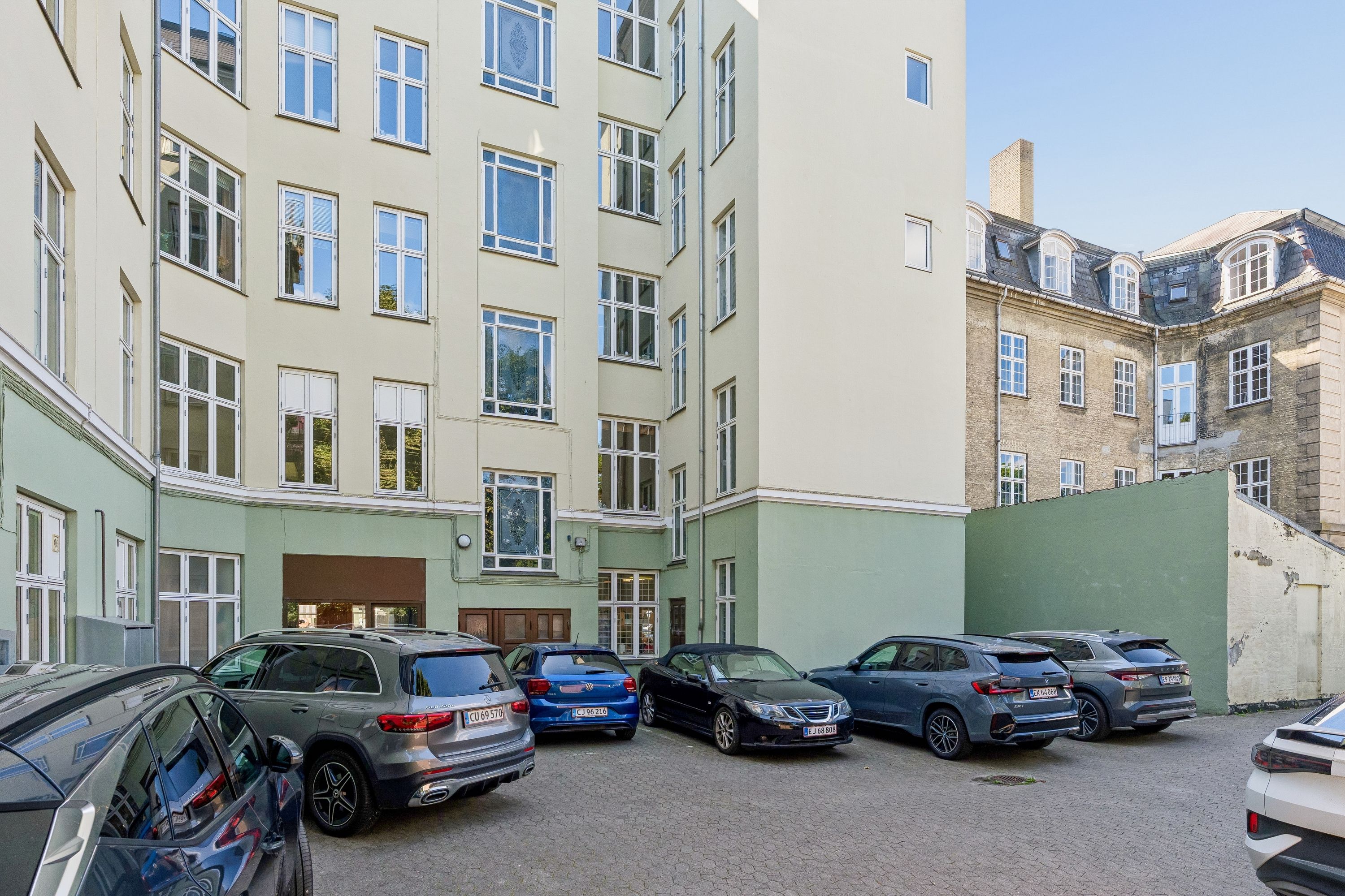 Flere biler parkeret i en lille parkeringsplads foran et fire etagers lejlighedsbygning med mange vinduer og grønne vægge