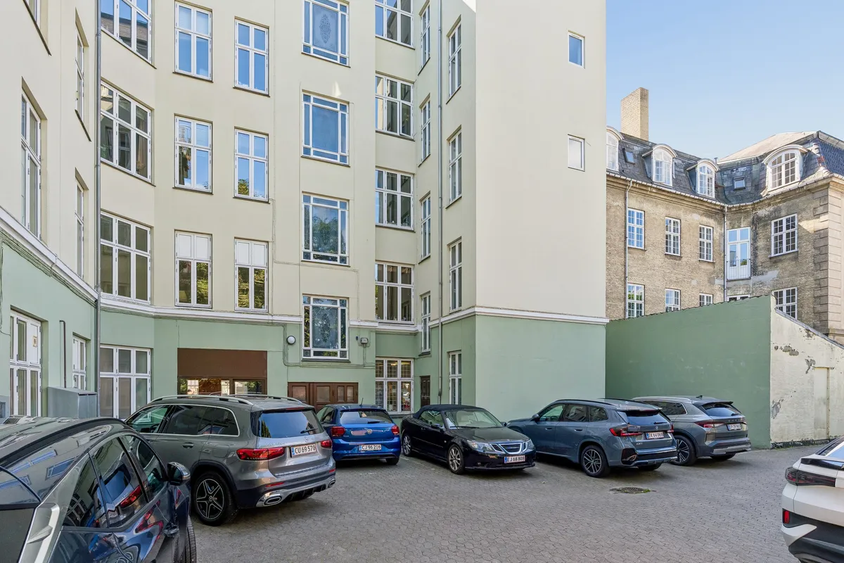 Flere biler parkeret i en lille parkeringsplads foran et fire etagers lejlighedsbygning med mange vinduer og grønne vægge