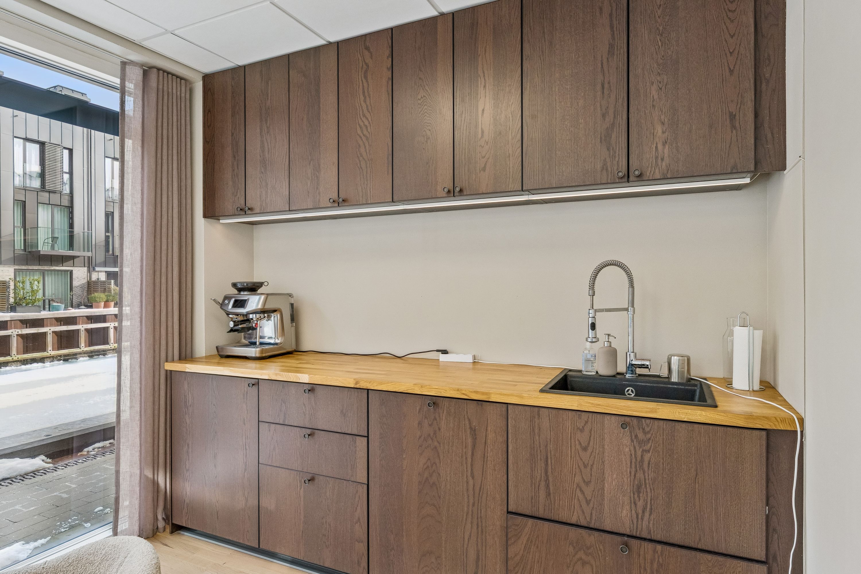 Kontor med lyse lofter og moderne møbler. Trægulv og en stor vinduesfront. Køkken med moderne apparater og træarbejde. Rørforsyning og skabe i mørkt træ.