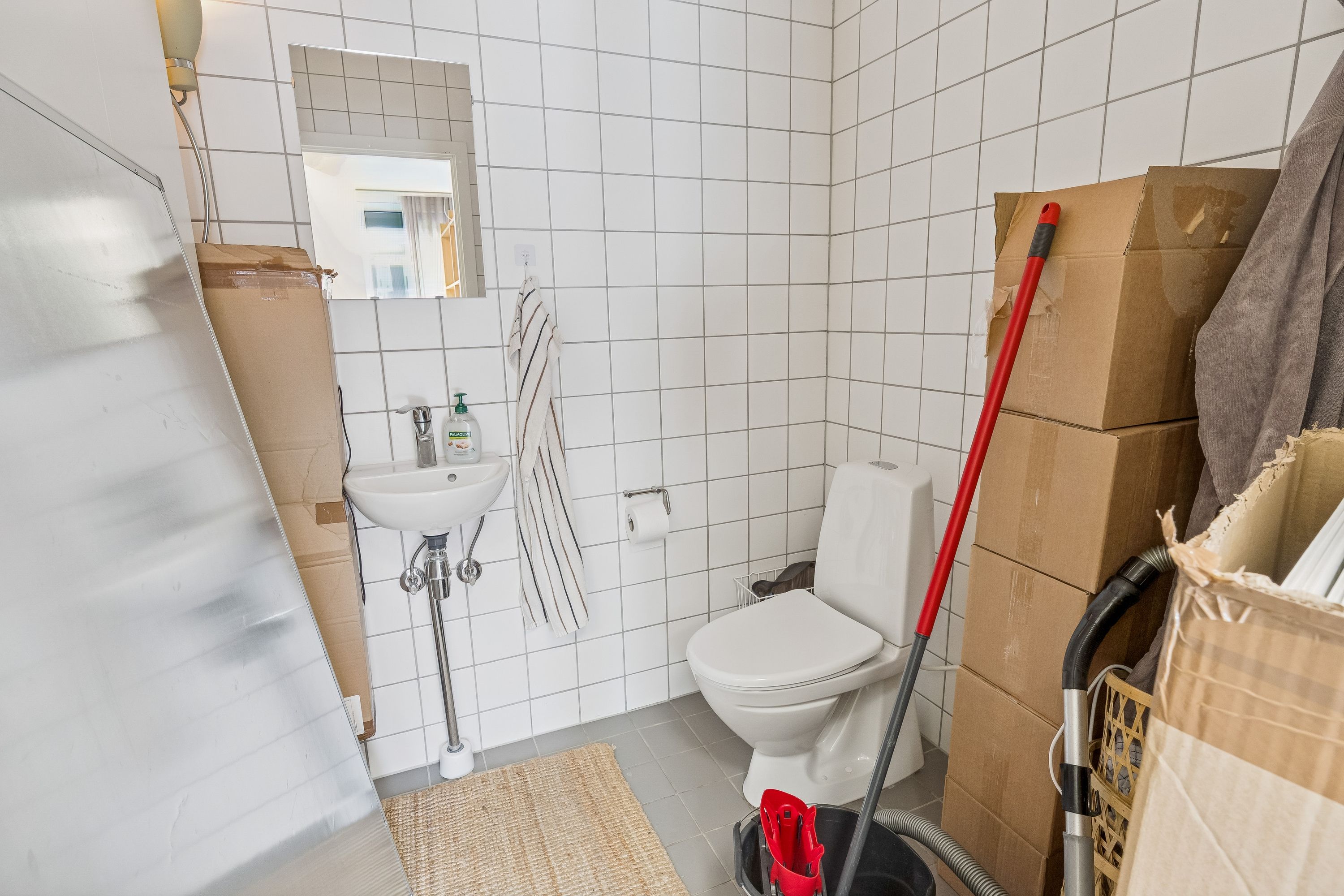 En hvid toilet i et lille badeværelse med et håndklæde og en sæbeflaske ved siden af. To papkasser står på gulvet og en støvsuger er lænet mod væggen.