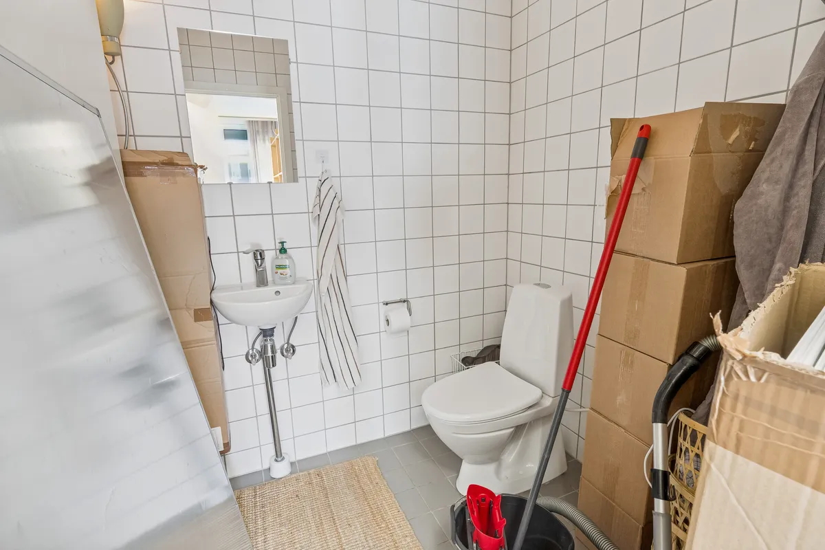 En hvid toilet i et lille badeværelse med et håndklæde og en sæbeflaske ved siden af. To papkasser står på gulvet og en støvsuger er lænet mod væggen.