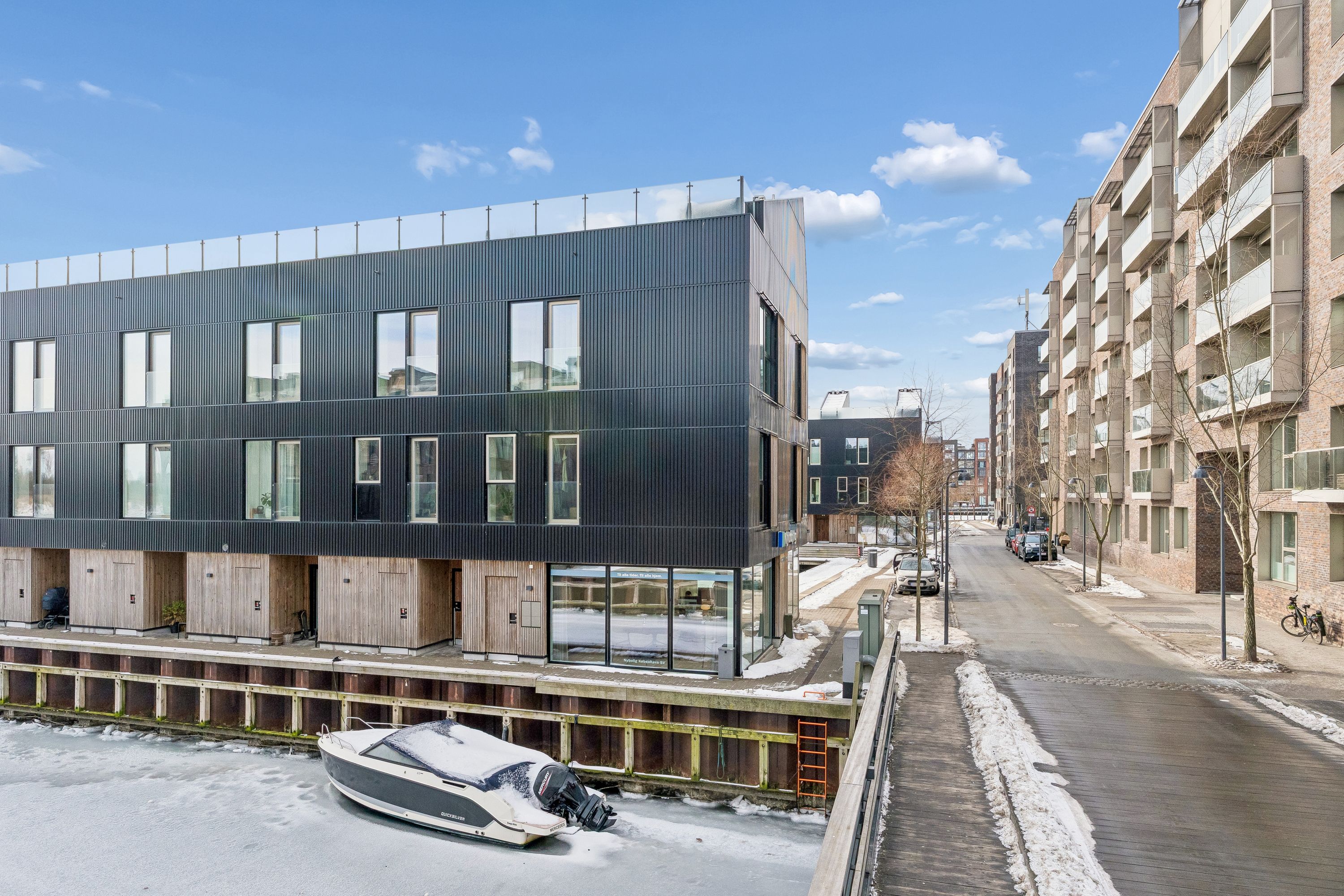 Moderne kontorbygning i sort og brun metal og glas med en række vinduer og balkoner. Vinterlandskab med sne og biler parkeret langs siden af en vej. En motorbåd er parkeret på en isdækket havn