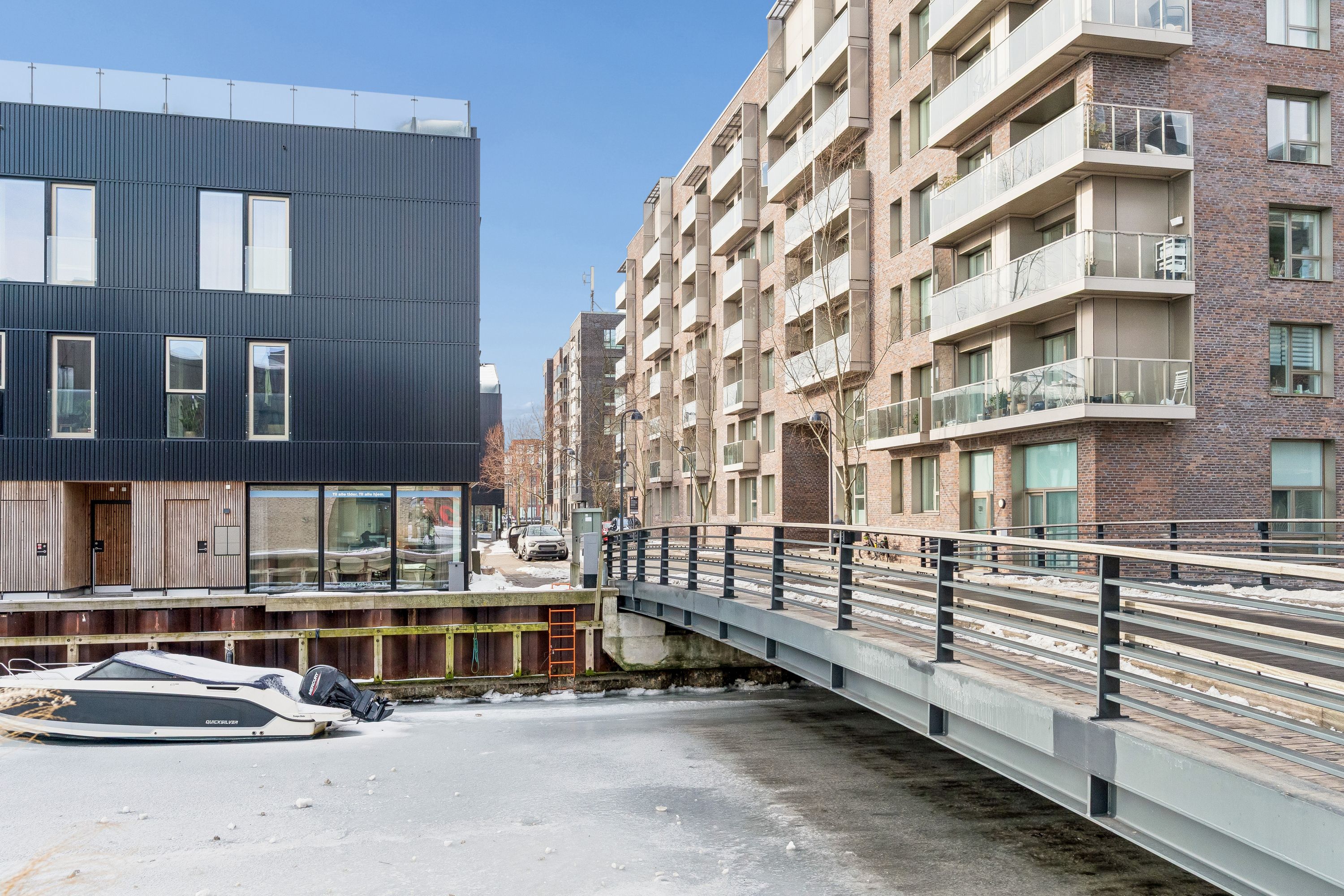 Lejlighedsbygning med glasfacade og balkoner og en båd parkeret ved kajen og en bro foran