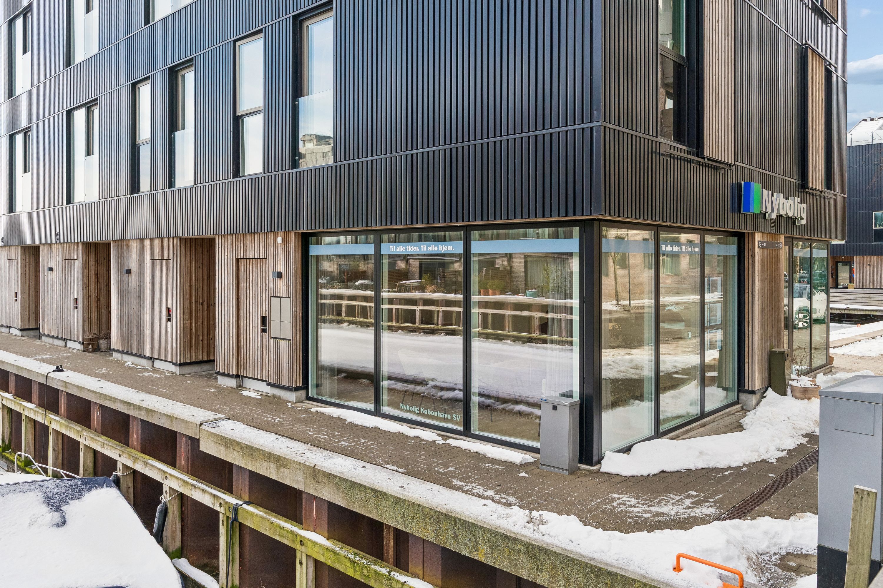 Moderne bygning med sort facade og store glasruder med Nybølle logoet og teksten "Til alle tider. Til alle hjem"