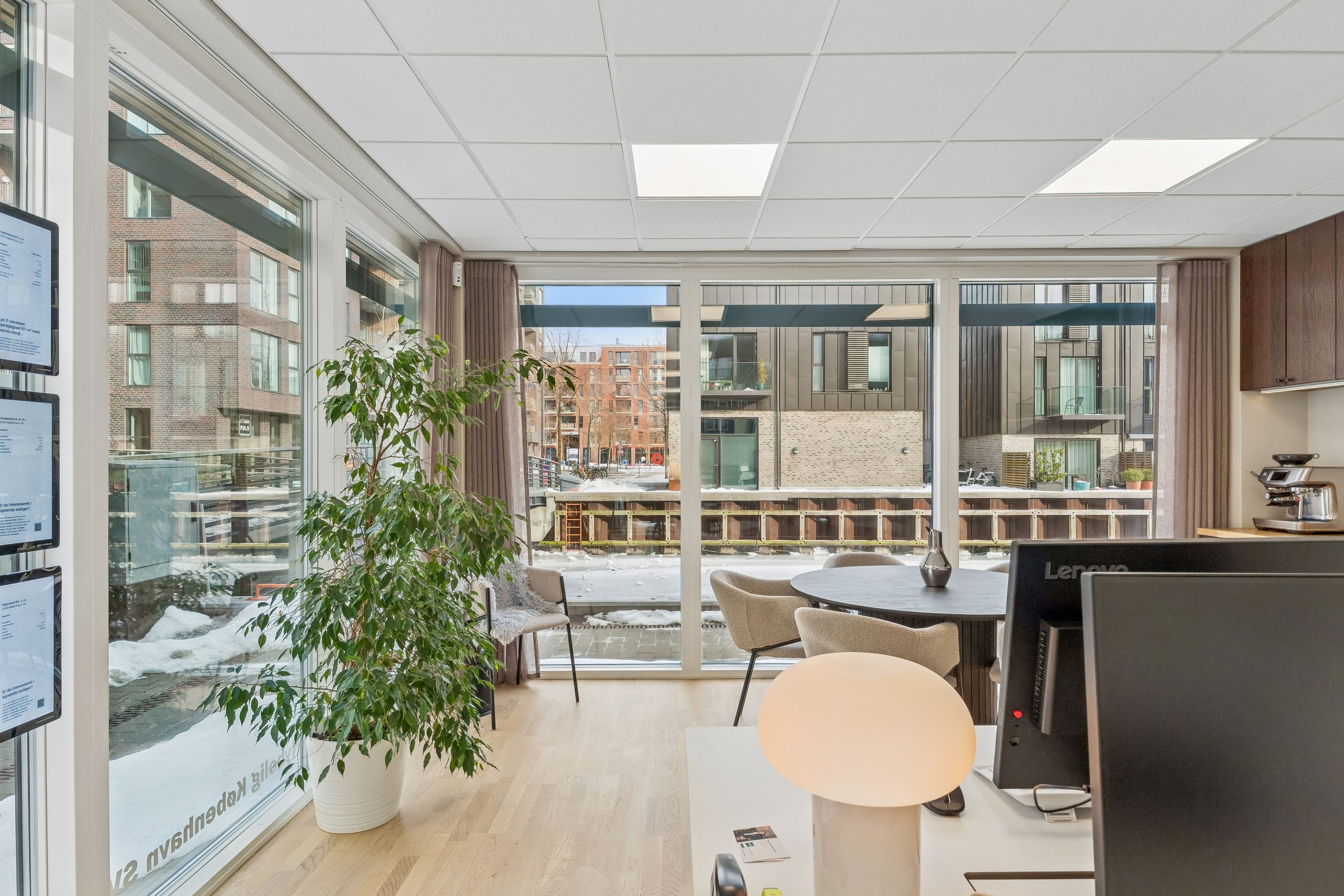 Kontor med smukke glasruder og et træ med en høj lampe i en hjørne. En rund bord med to stole og en monitor.