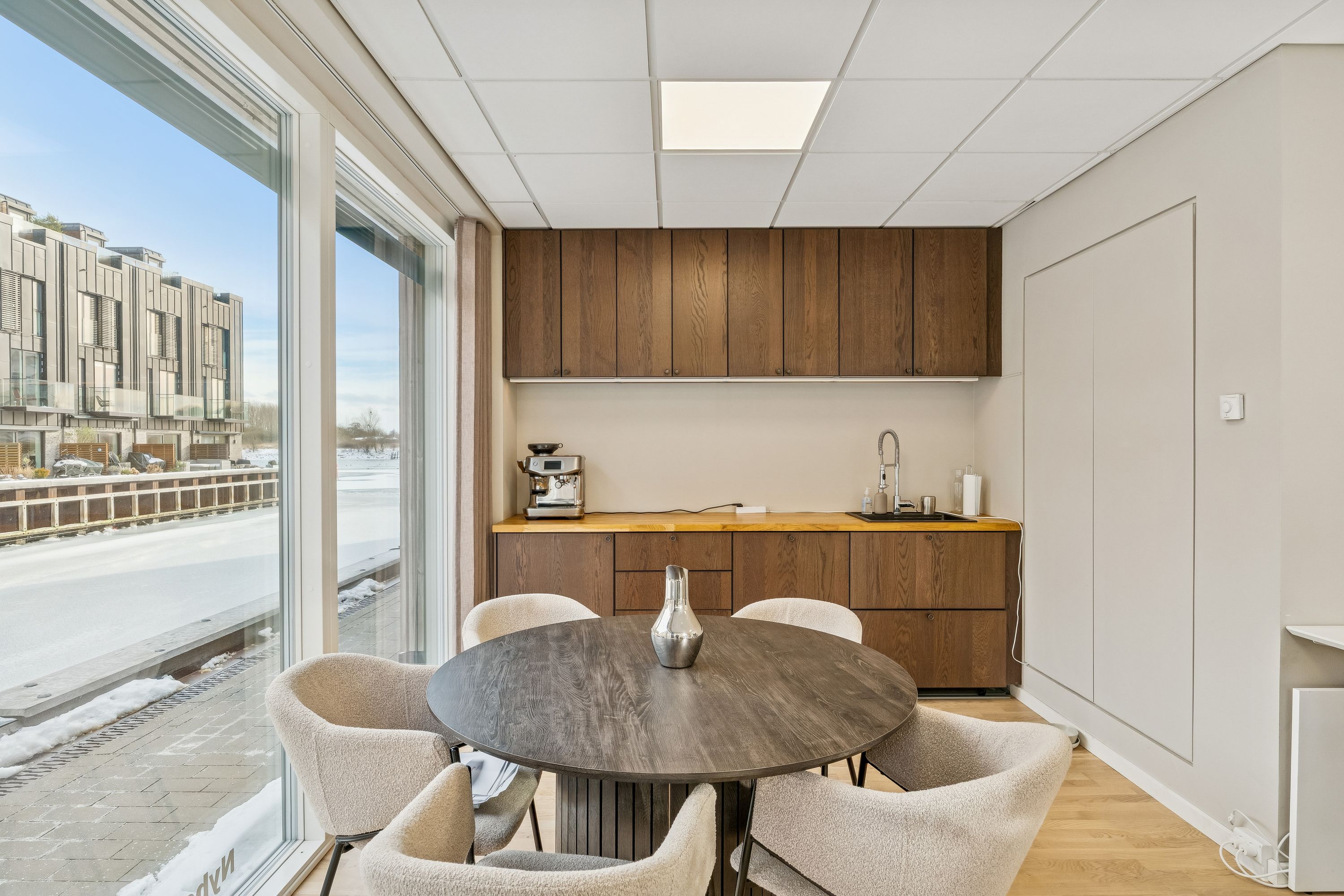 Kontor med runde bord og stole samt kaffemaskine og vask. En stor glasvindue med udsigt til sne og bygninger. Lys beige vægge og træfronter på skabe og skrivebord