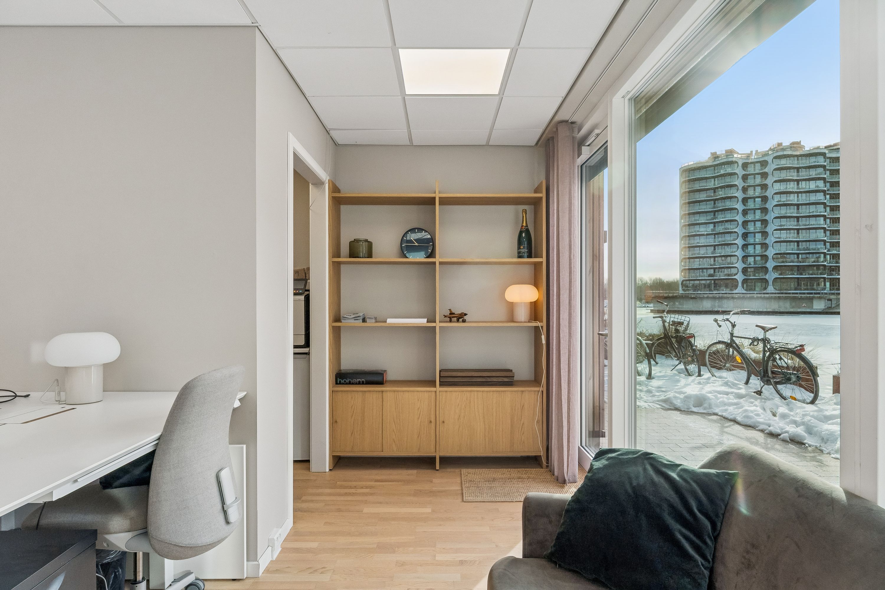Kontor med højloft og stort glasvindu, der vender ud mod en isdækket havn med cykler. Vinduet skuer ud på bygninger og sne på en bred flod.