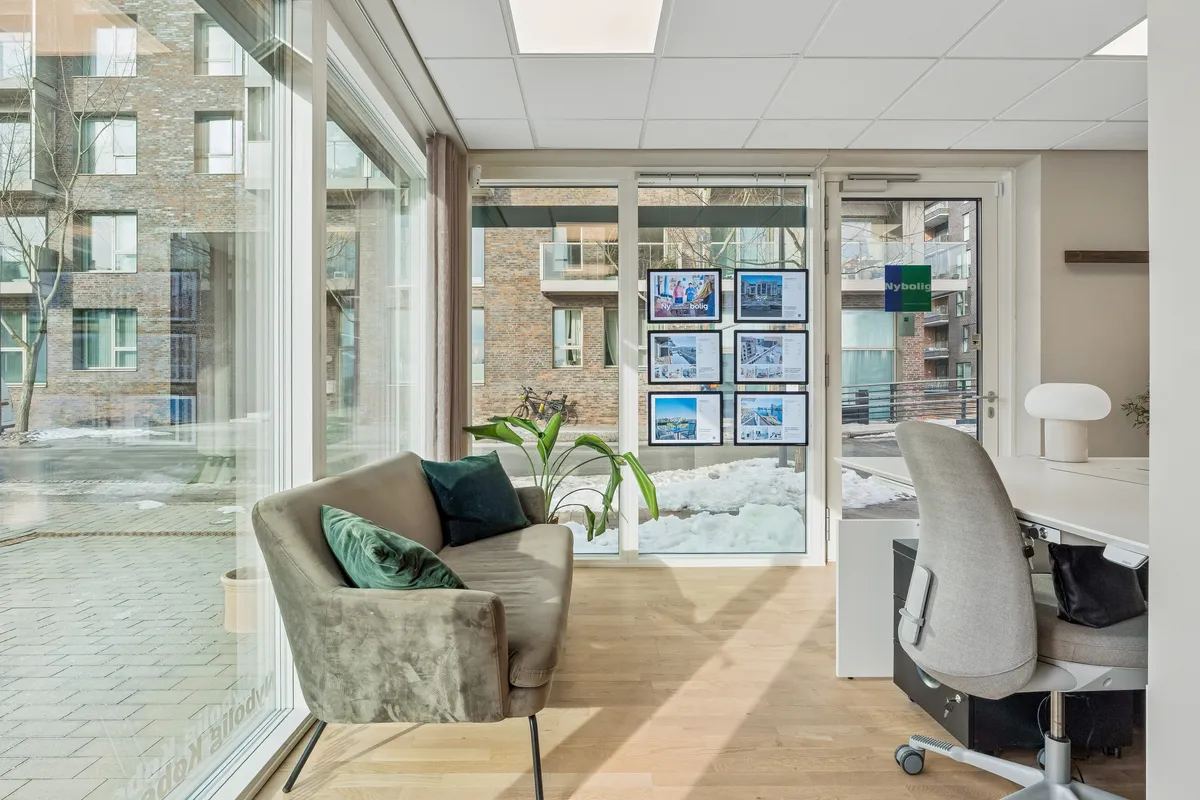 Lys og moderne kontor med glasvinduer, stol, bord, sofa og planter samt udsigt til byggede boligblokke med sne og cykel.