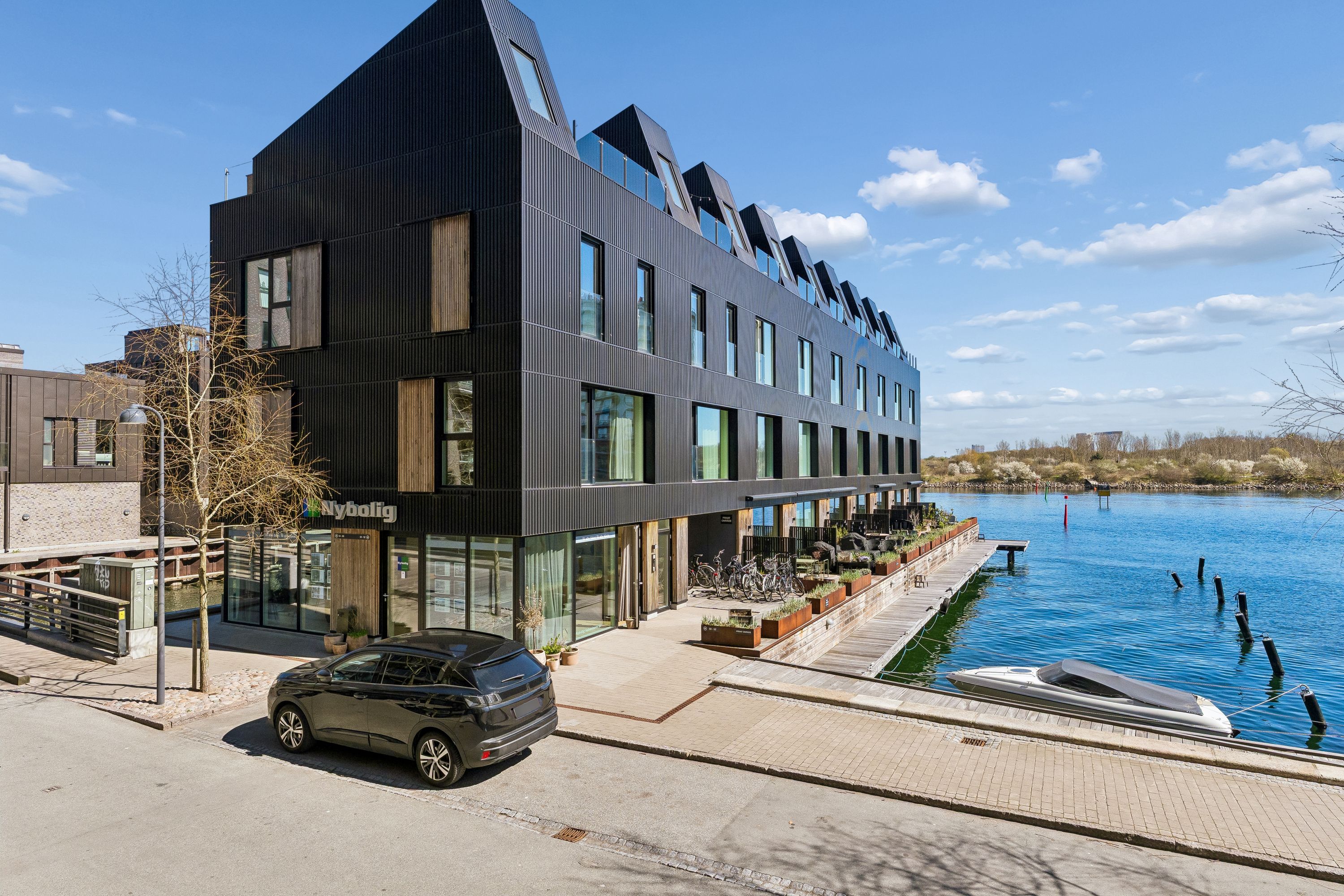 Store bygning med moderne design og glasfacade ved vandkanten. Lejemål med parkerede biler, udsigt til vandet og træer. Alle elementer er i perfekt stand og bygningen er sammensat af sort og brun materiale.