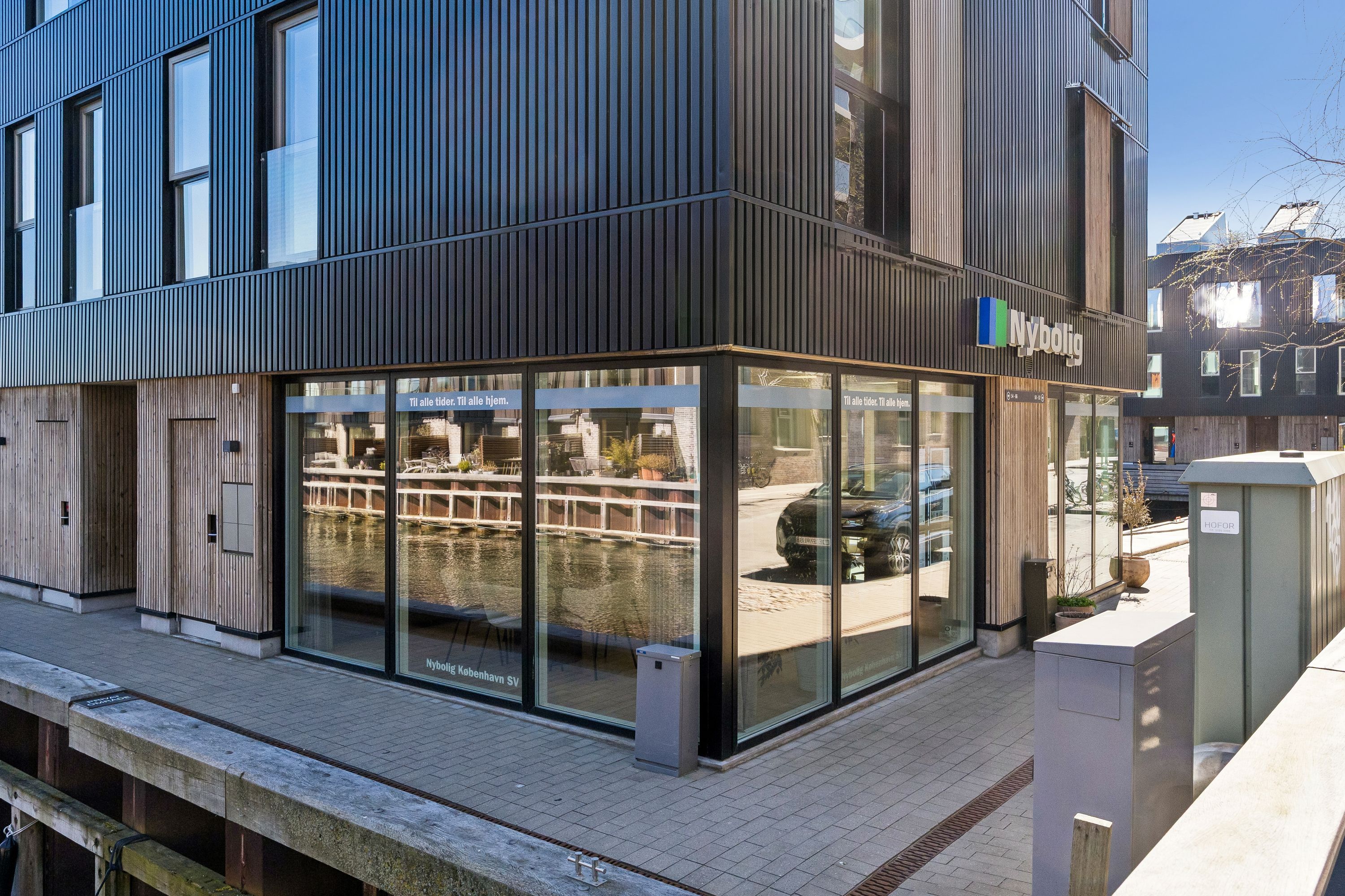 Nybolig butik til leje på Teglholmens Østkaj 50 i København SV med store glasvinduer og moderne facade i sort og brun. Bilparkering foran og udsigt over vandløb.