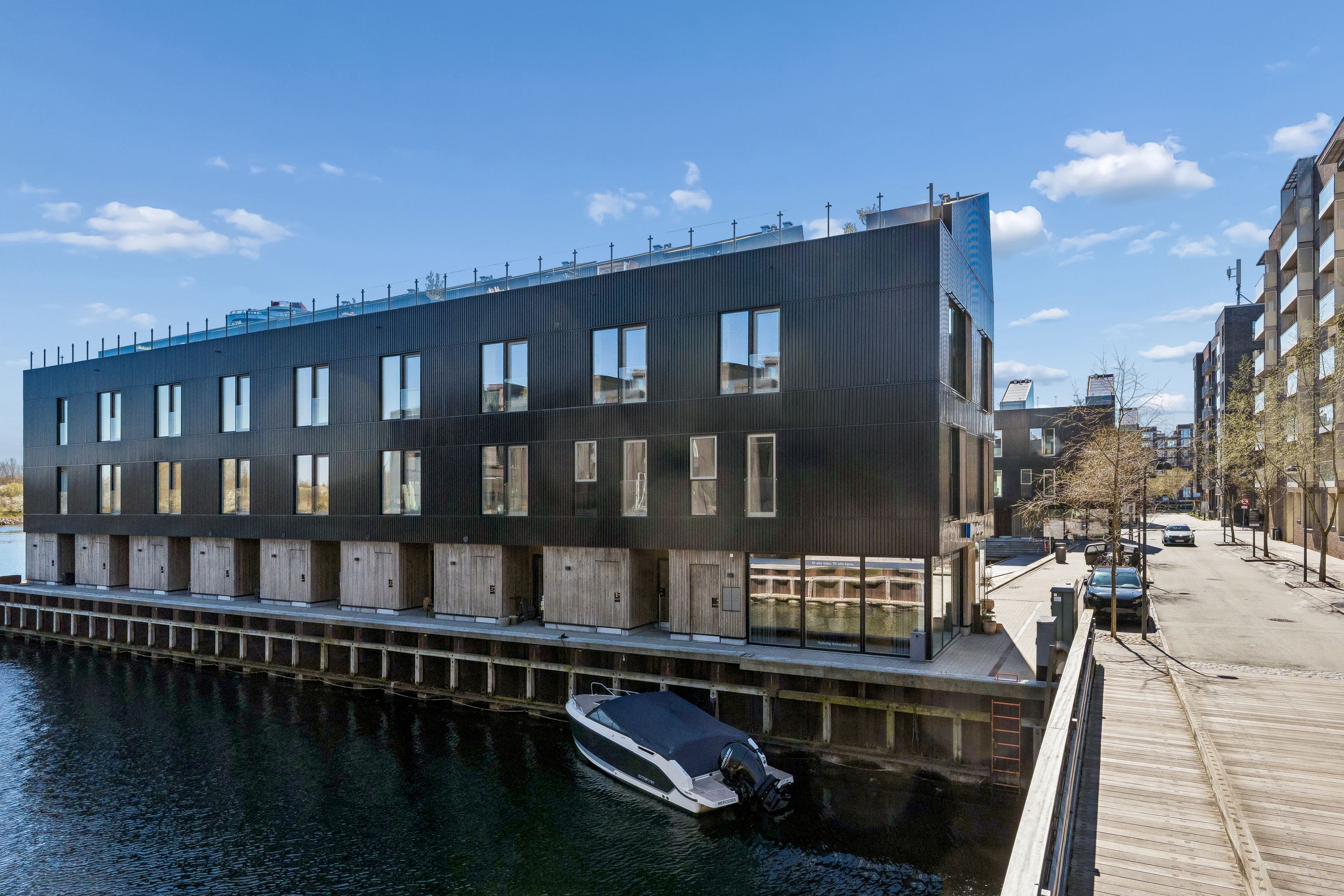 Moderne bygning med glasvinduer og træelementer langs vandkanten, et parkeret båd, træbro og en bil parkeret på gaden