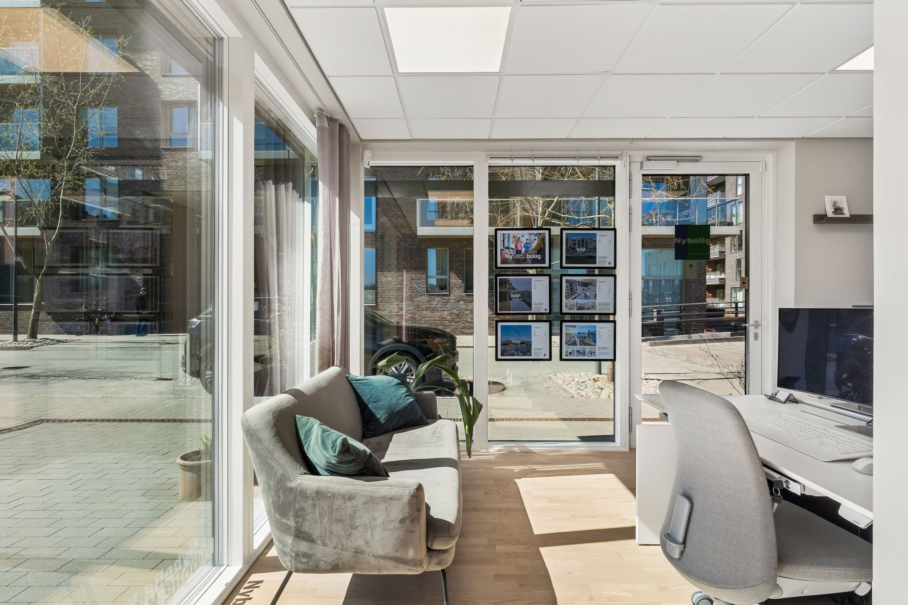 Moderne stue med skrivebord og stol, sofa med pude og eksempler på udlejningsboliger på væggen, glasvinduer med udsigt til gaden og bygninger