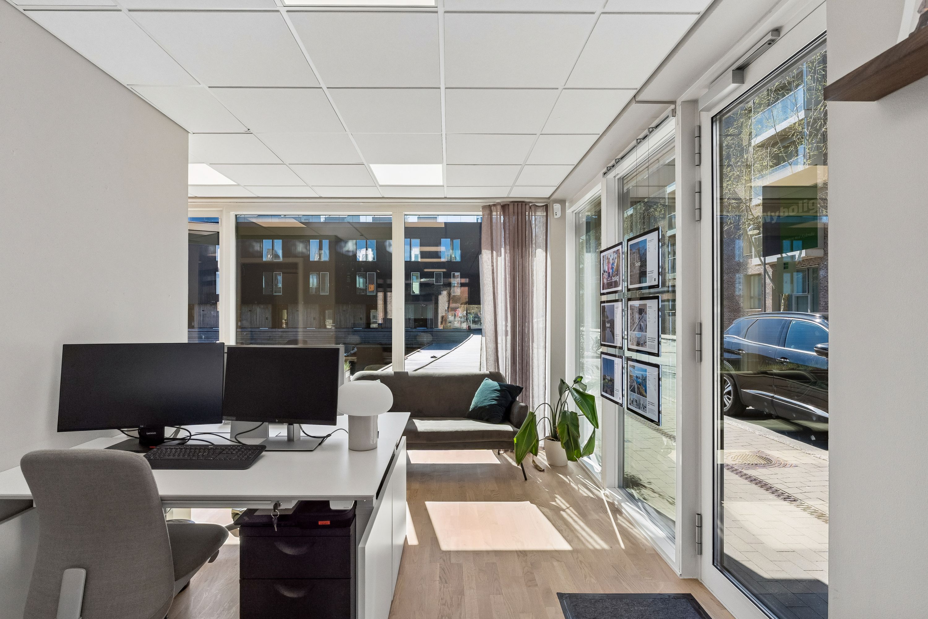 Kontor med dobbeltskærm, skrivebord, stol, plante, sollys, gulv, vinduer, køretøj og reklame.