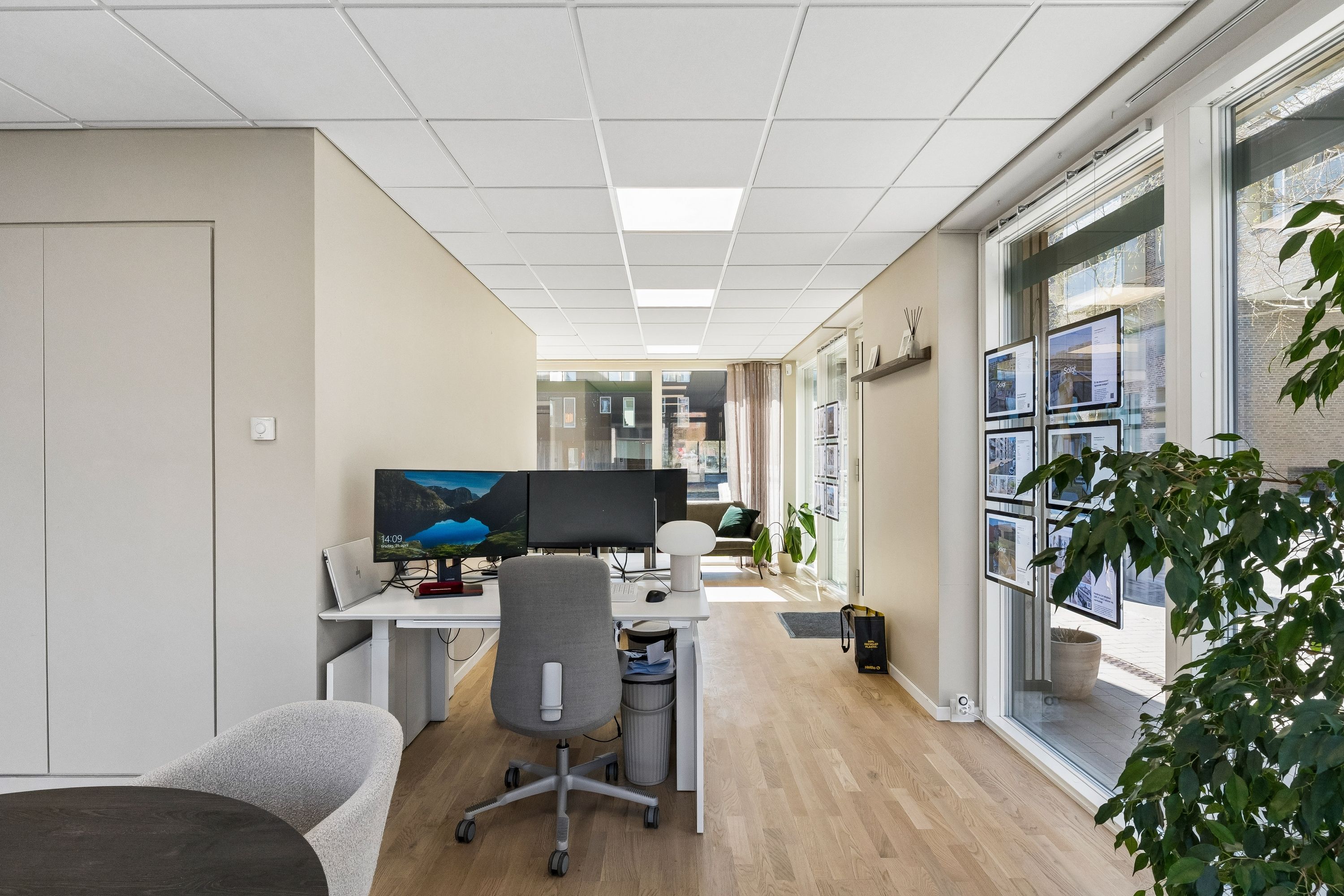 Kontor med højt loft og lyse loftslamper, skrivebord med to skærme og kontorstol, glasdør og udsigt til grønne planter og udsigt til bygninger udenfor