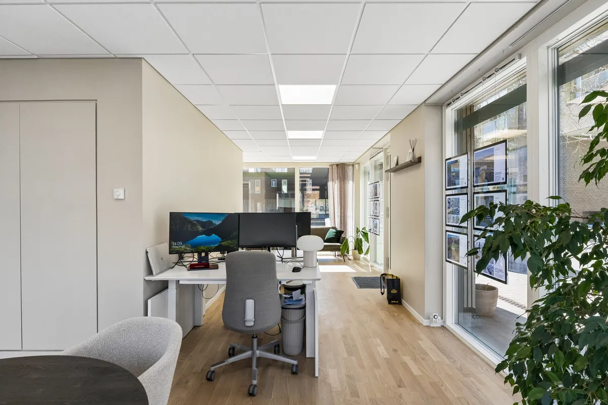 Kontor med højt loft og lyse loftslamper, skrivebord med to skærme og kontorstol, glasdør og udsigt til grønne planter og udsigt til bygninger udenfor